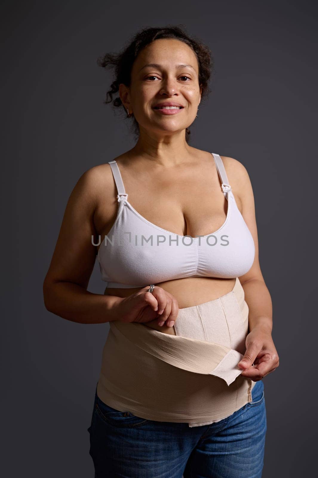 Authentic studio portrait of multi ethnic smiling woman, young mother putting on elastic bandage after cesarean c-section, looking at camera, isolated gray studio backdrop. Skin and body care concept