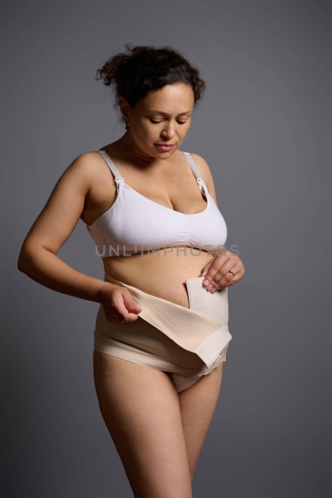 Vertical portrait of Latin American middle aged naked woman, young mother in underwear after c-section, wearing elastic bandage, isolated over gray studio background. Postpartum. Post pregnancy time