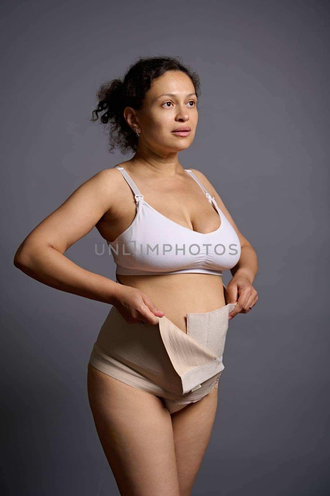 Multi ethnic woman, young mother putting on a bandage after c-section, looking away, isolated on gray studio background by artgf