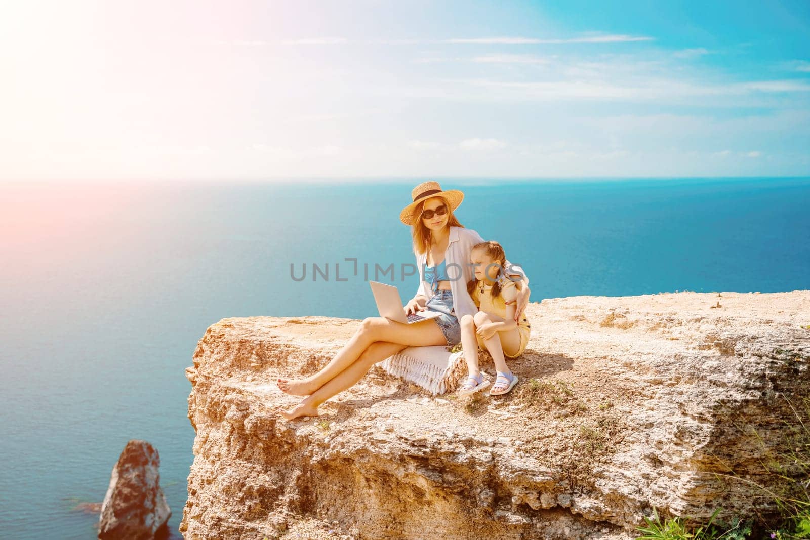 Freelance woman with her daughter working on a laptop by the sea, typing on the keyboard, enjoying the beautiful view, highlighting the idea of remote work. by Matiunina