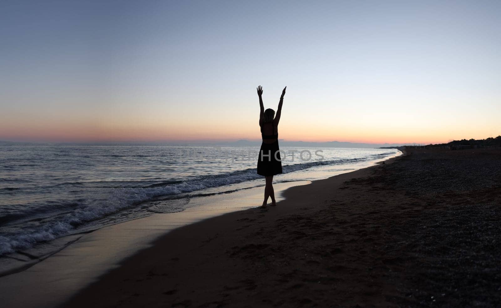 Woman with her hands up on beach at sunset back view. Happiness and outdoor recreation concept