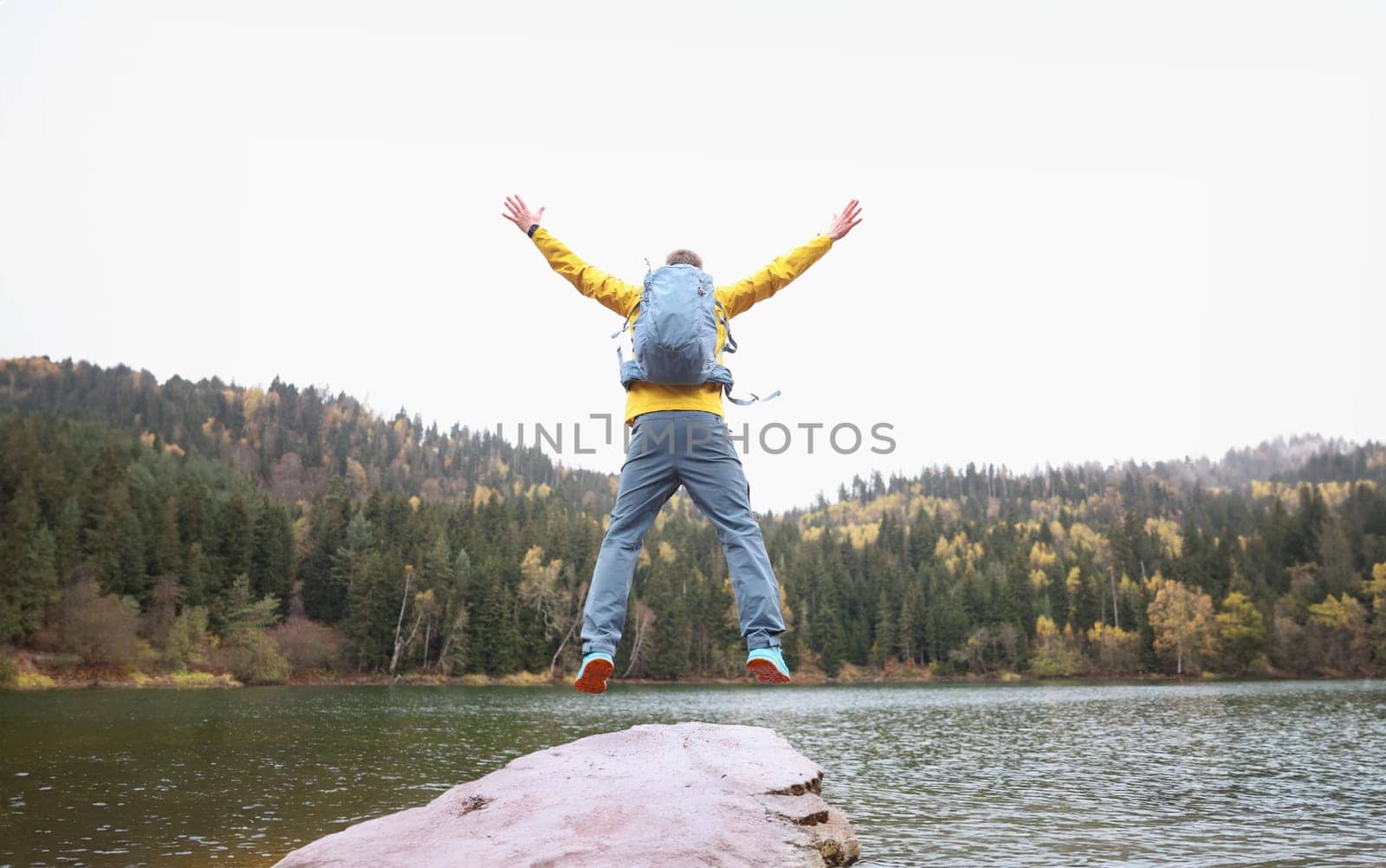 Happy male traveler with backpack jumping near forest lake while reaching goal back view closeup by kuprevich