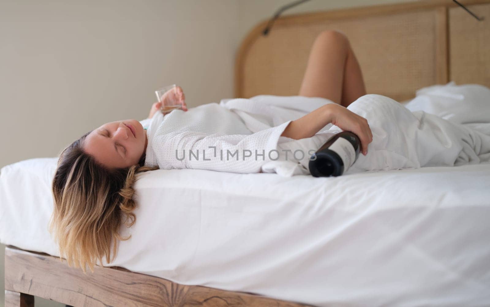 Drunk woman in bath robe lying in bed with bottle of wine in hotel by kuprevich