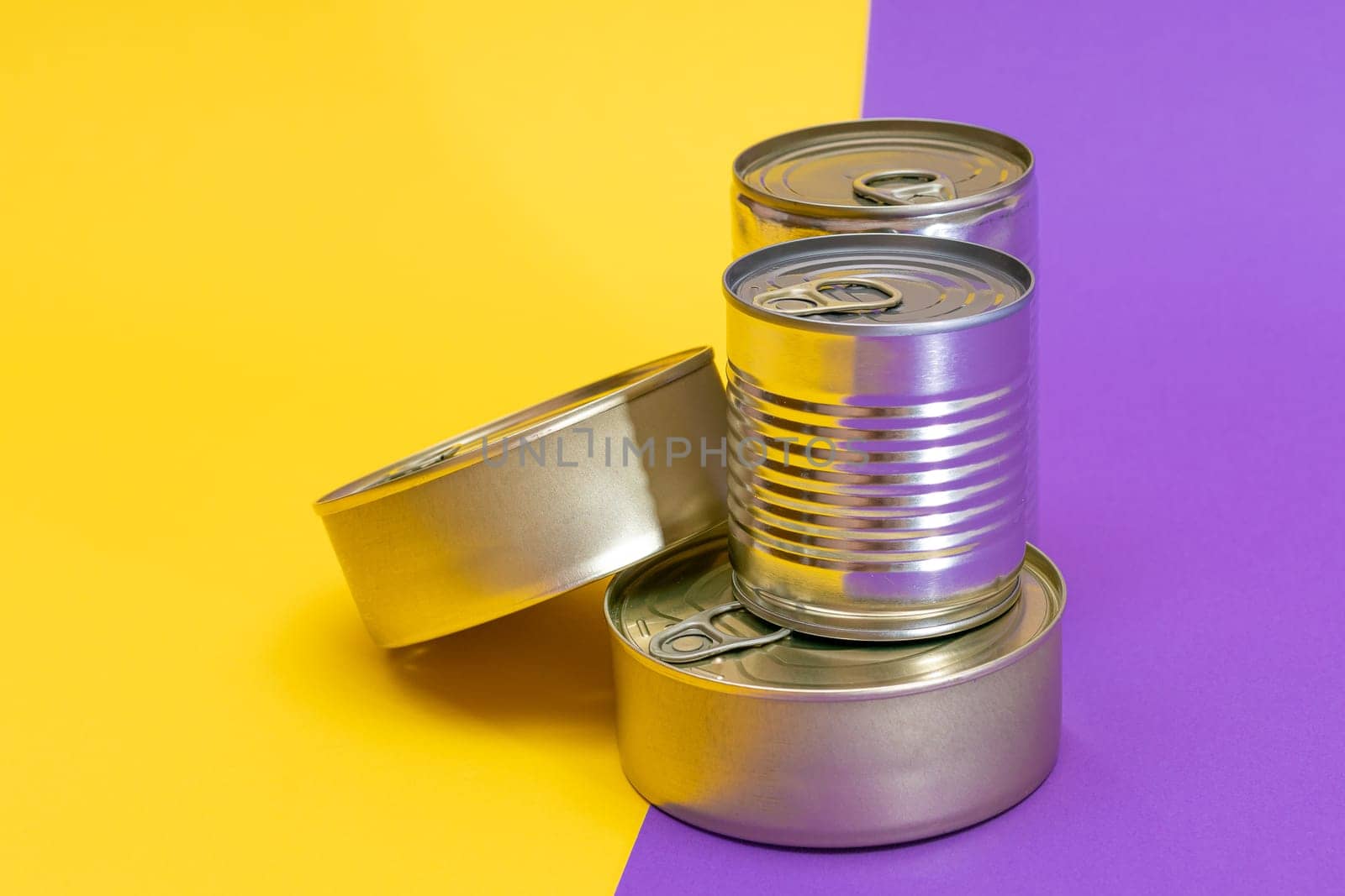 A Group of Stacked Tin Cans with Blank Edges on Split Yellow and Violet Background. Canned Food. Different Aluminum Cans for Safe and Long Term Storage of Food. Steel Sealed Food Storage Containers