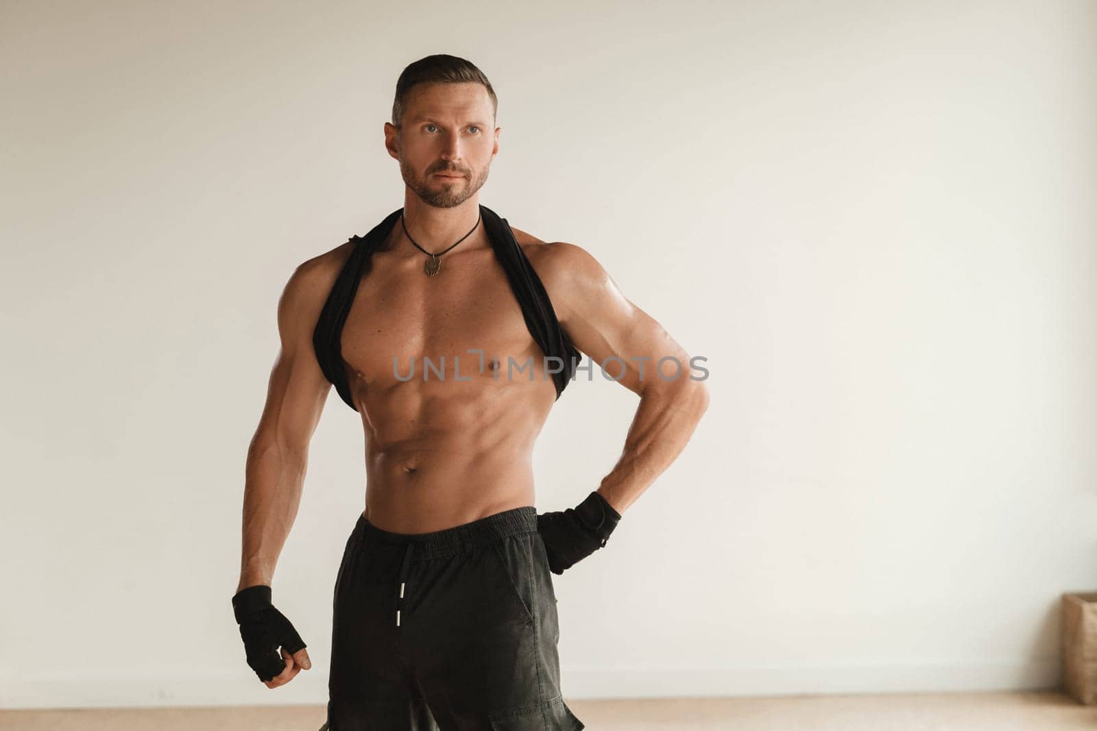 An adult muscular man with a naked torso stands in a room on a light background.