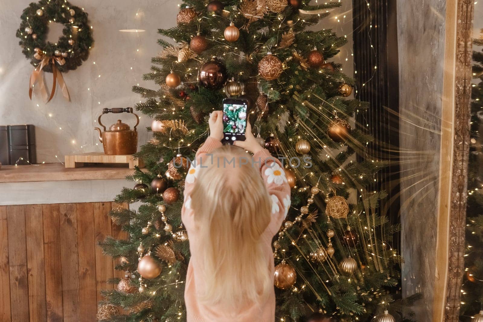 A little blonde girl photographs balloons on a Christmas tree in a festive interior decorated in a New Year's style. The concept of a merry Christmas