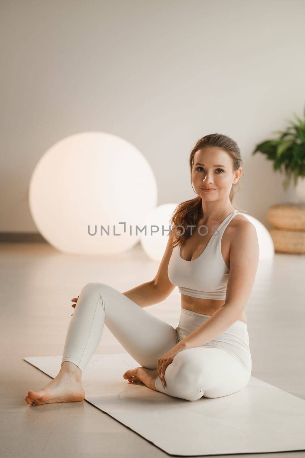 Portrait of a girl in white clothes sitting on a mat before doing Yoga indoors by Lobachad