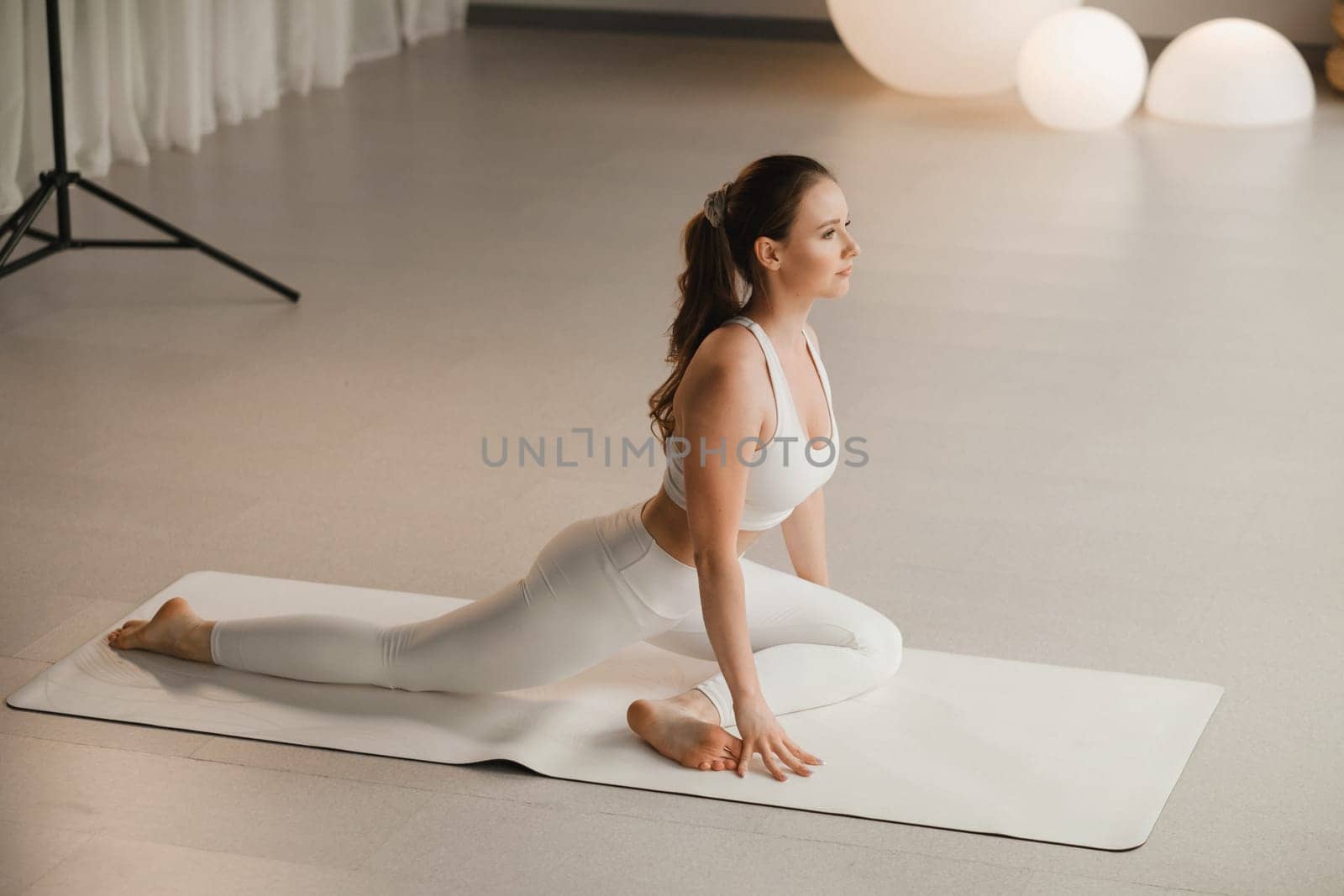 A girl in white clothes does yoga on a mat indoors by Lobachad