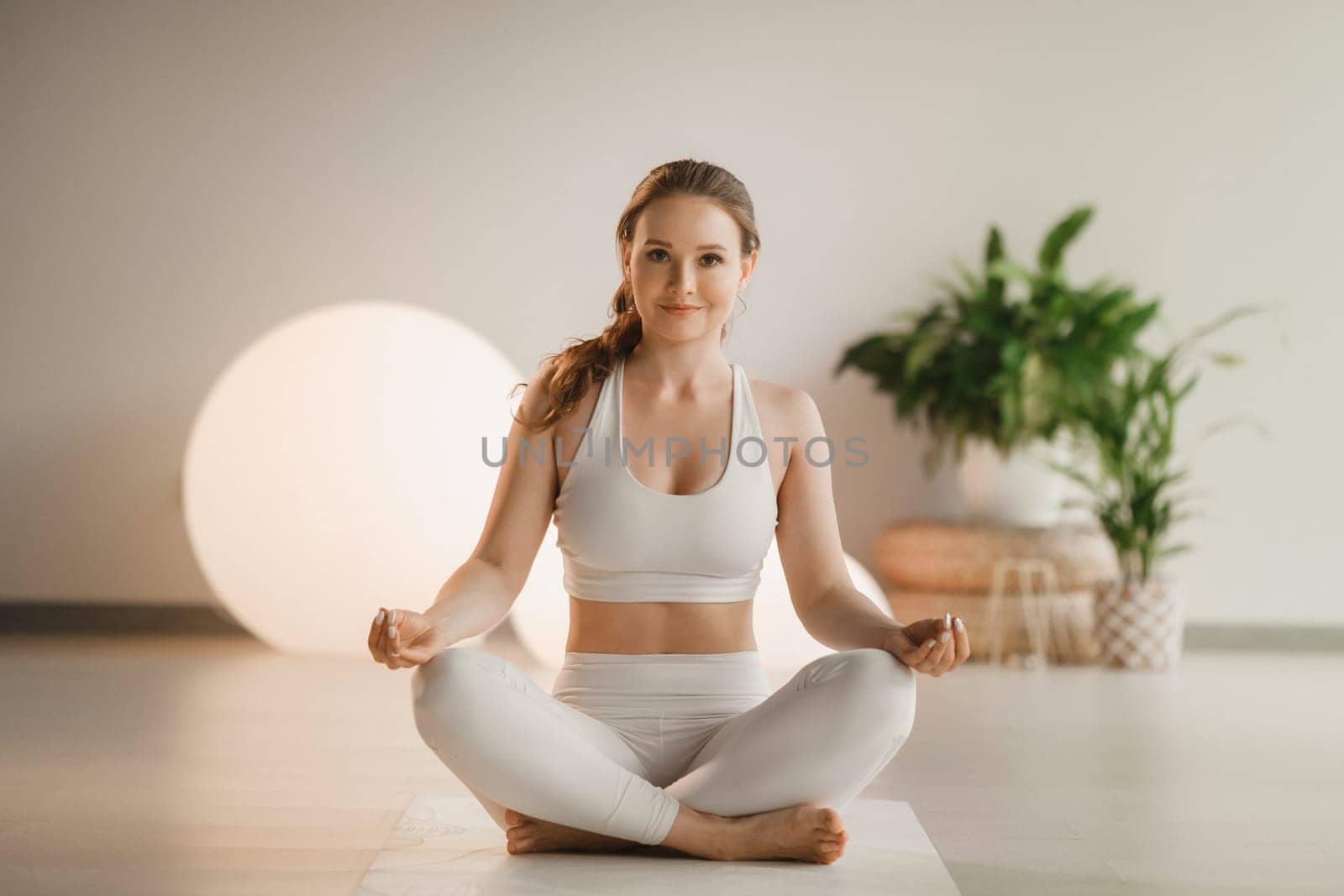 Portrait of a girl in white clothes sitting in a lotus position on a mat at an indoor yoga class.