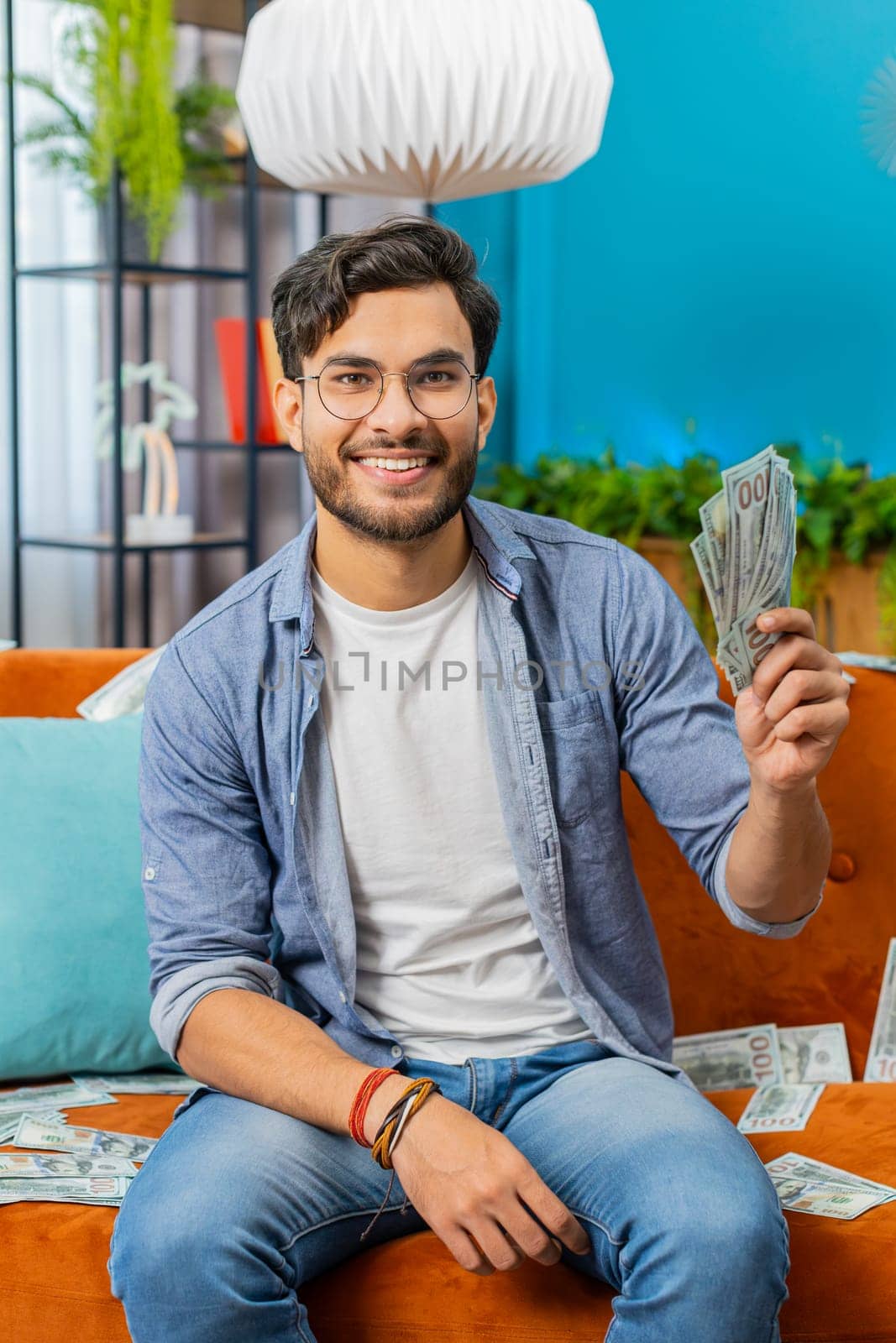 Successful happy rich winner Indian man counting money cheering up with high profits, lottery game win, satisfied of wealth income earnings salary on home couch full of dollar cash banknotes. Vertical