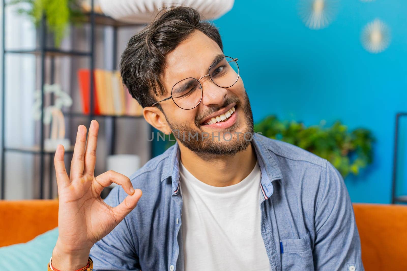 Okay. Happy cheerful Indian man looking approvingly at camera showing ok gesture, positive like sign, approve something good in living room sitting on sofa. Arabian Hindu guy at modern home apartment