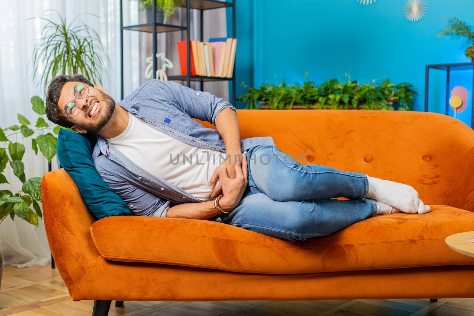 Indian young man lying on sofa feeling sudden strong abdominal stomach ache gastritis problem. Bearded Arabian guy having symptom poisoning diarrhea indigestion peptic ulcer pancreatitis at home room