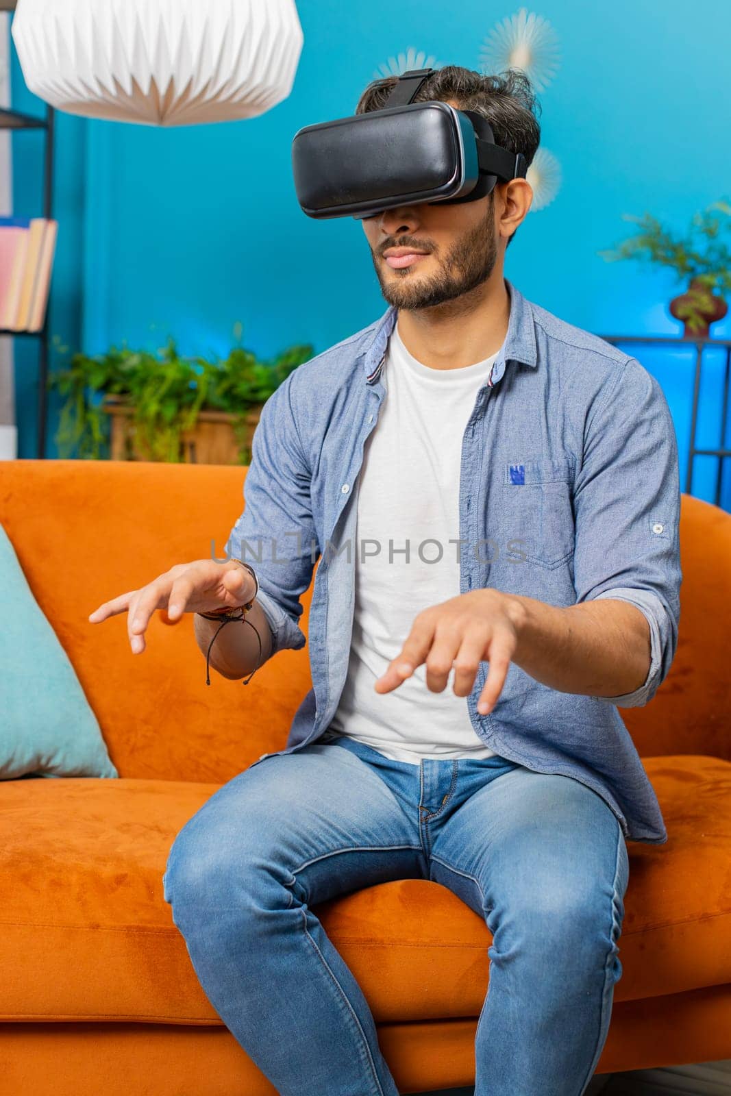 Indian man using virtual reality futuristic technology VR app headset helmet working typing on virtual keyboard, play simulation 3D 360 online video game at home apartment. Guy in goggles on couch