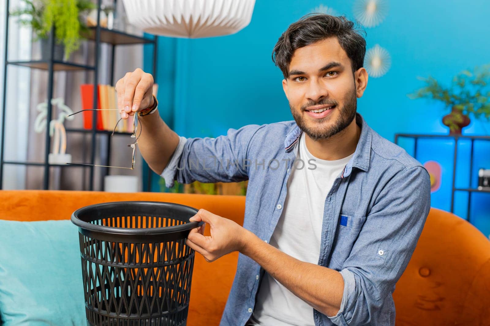 Happy Indian man taking off throwing out glasses into bin after medical vision laser treatment therapy surgery looking smiling at camera, heal, cure. Arabian guy sits on couch at home room apartment