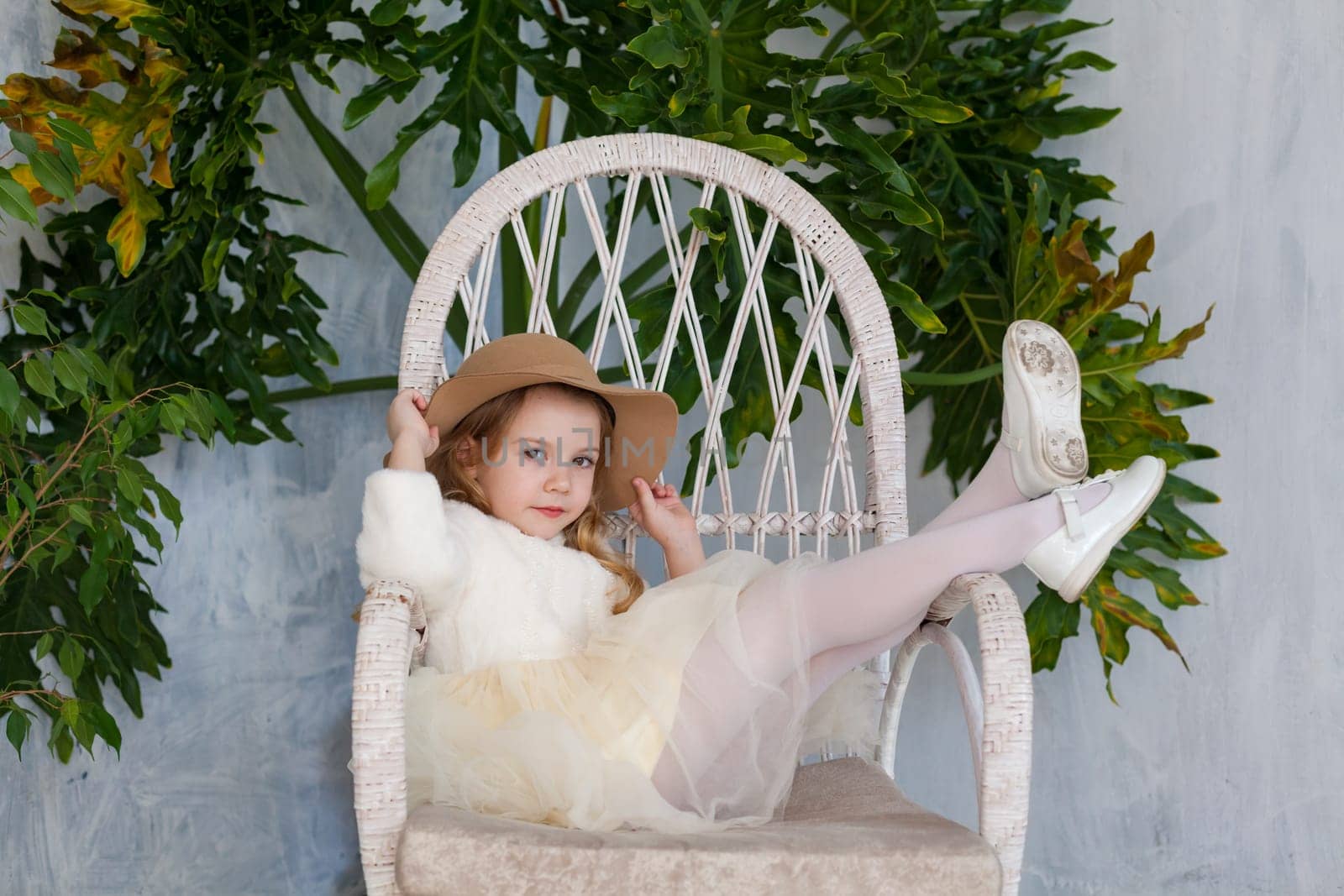 Little fashionable girl in a hat sits in a white chair by Simakov