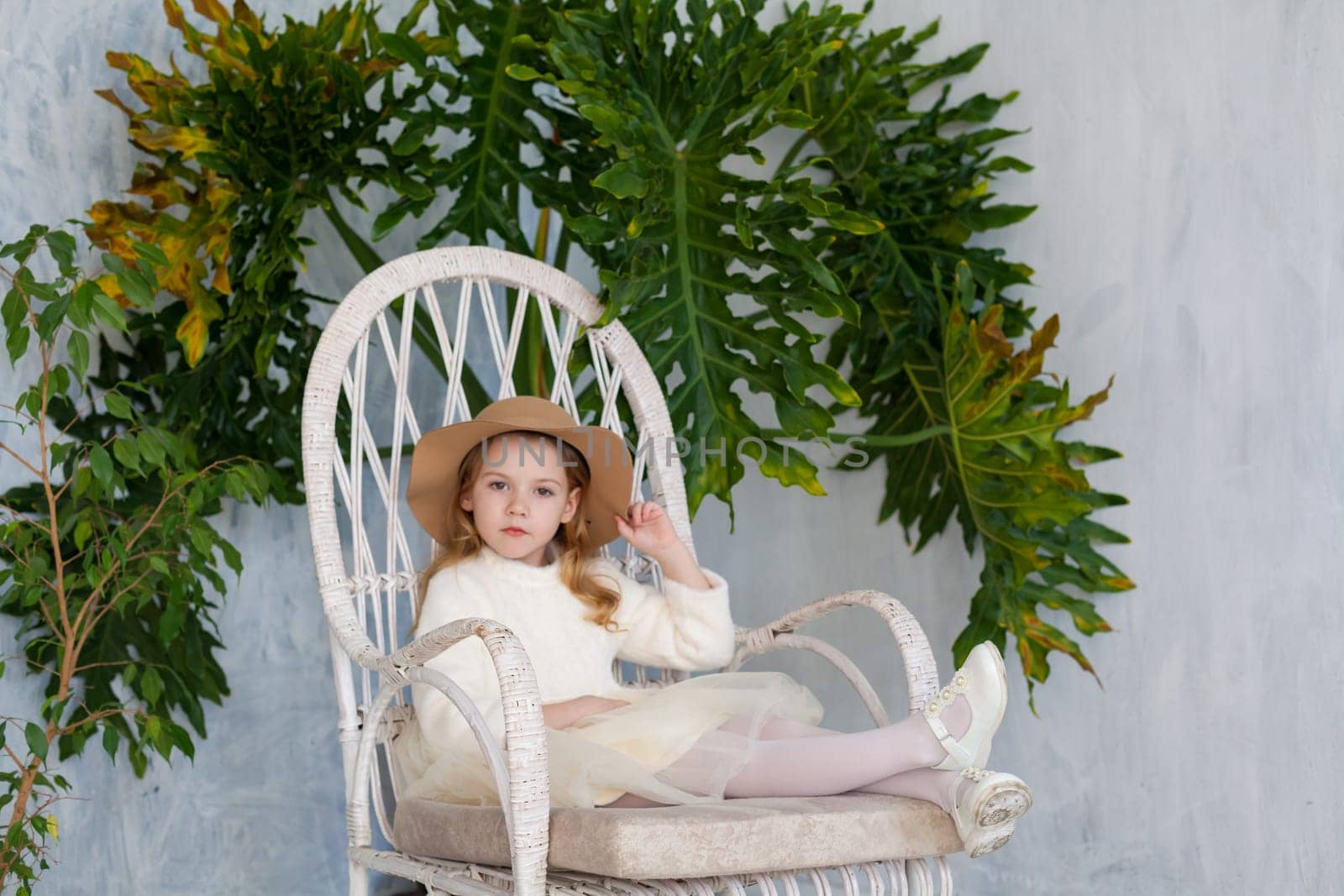 Little fashionable girl in a hat sits in a white chair by Simakov