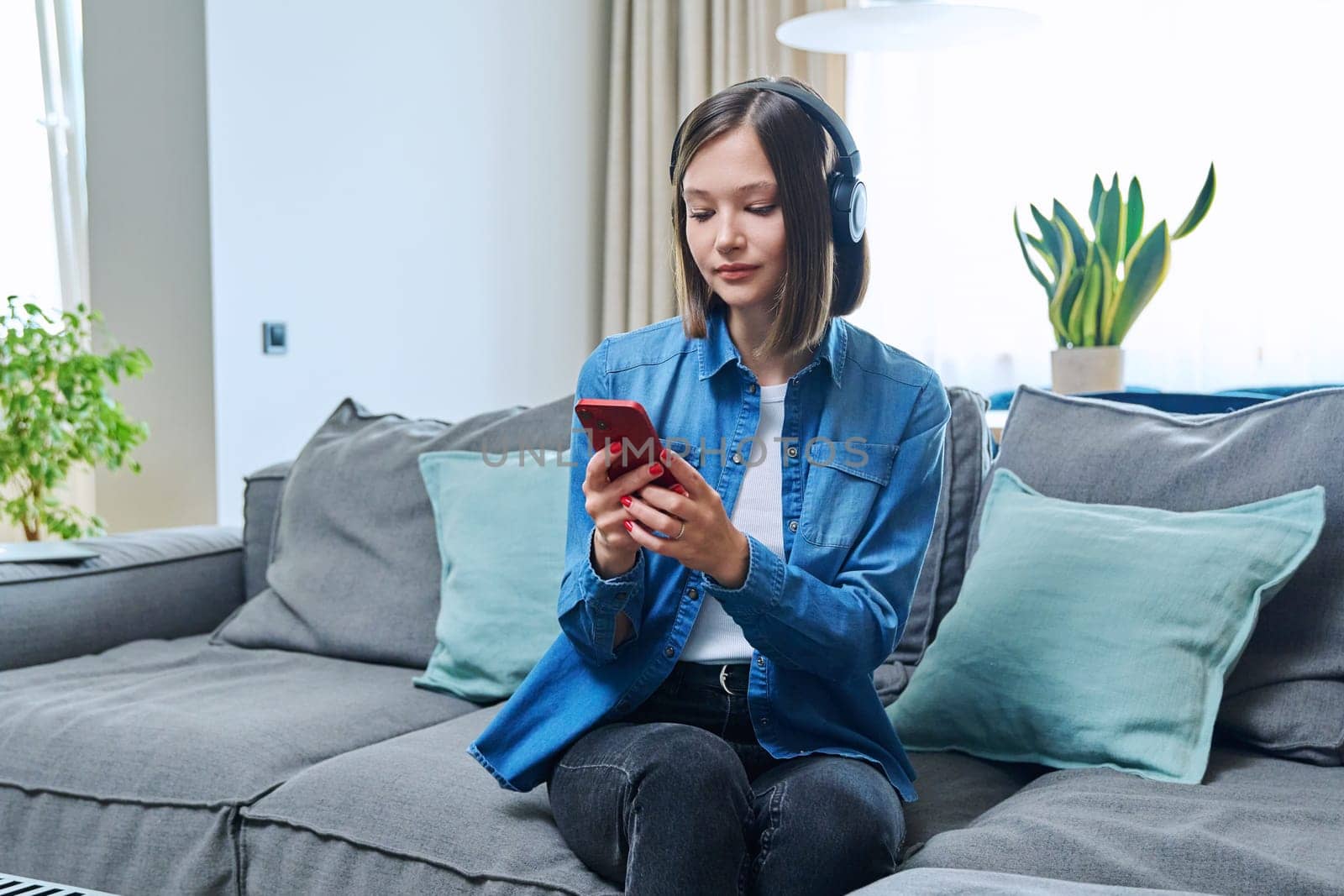 Young woman sitting on sofa in living room wearing headphones using smartphone, happy relaxing female listening music audio book learning languages chatting with friends, 20s student studying online