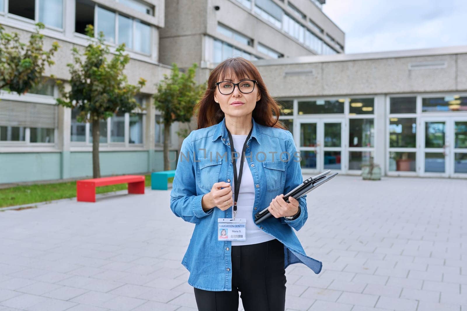 Middle-aged confident woman school teacher, mentor, pedagogue, psychologist, counselor, social worker with digital tablet in hands posing near school building, outdoor