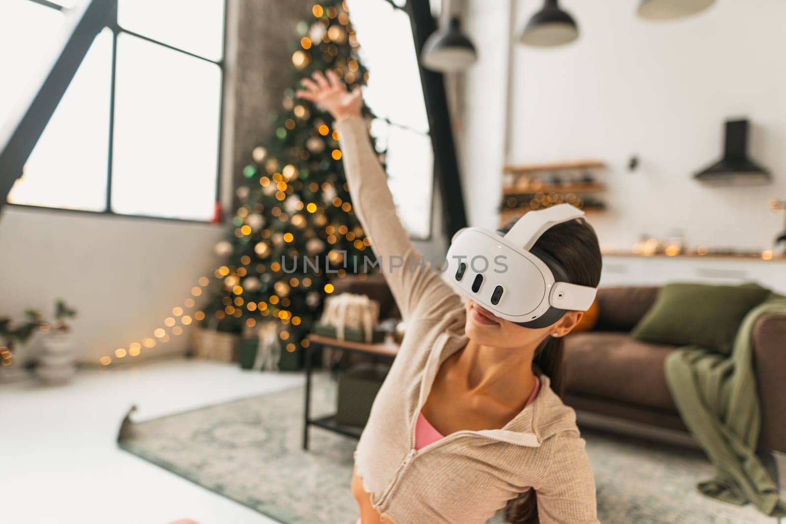 A fitness-oriented young woman practices yoga poses while wearing VR glasses by a Christmas tree. High quality photo