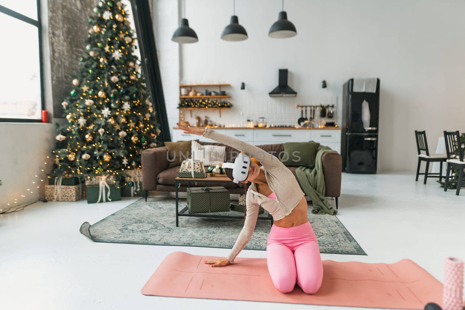 A lovely young woman is stretching in front of a Christmas tree while wearing virtual reality glasses. by teksomolika