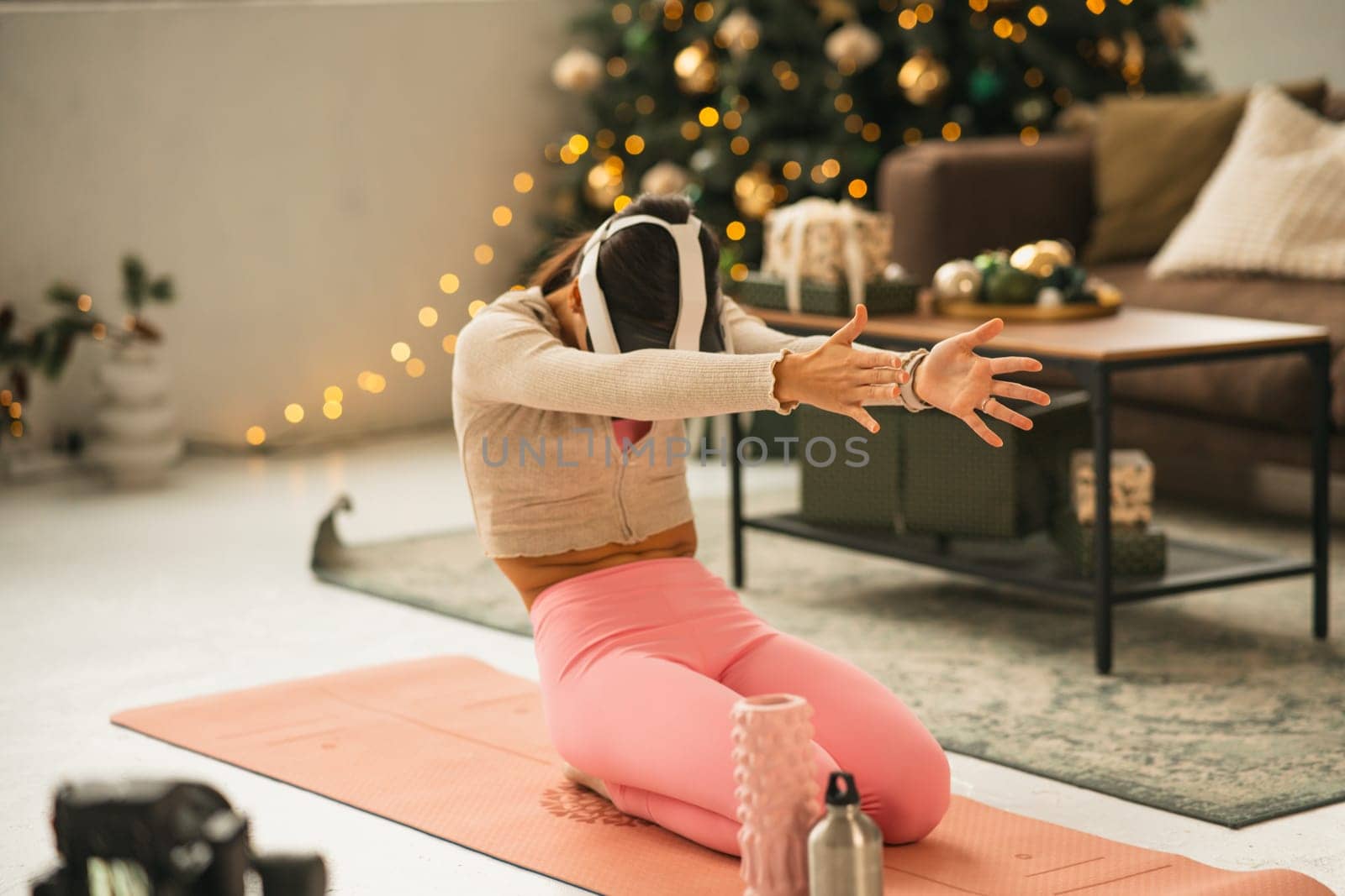 A fashionable young woman in sports attire, sporting a virtual reality headset, does yoga with a Christmas tree in the background. High quality photo