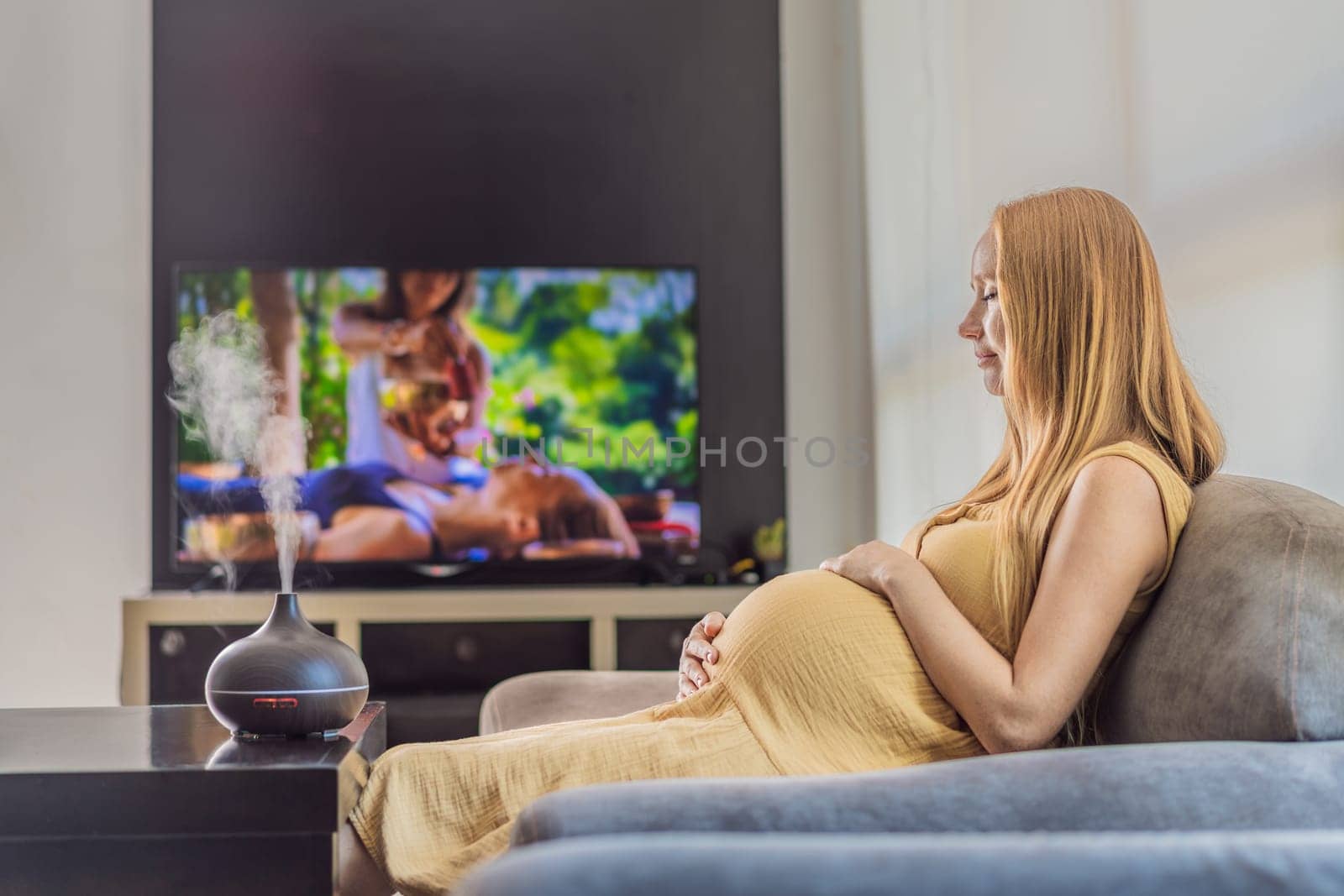 A blissful pregnant woman immerses in relaxation, savoring the soothing aroma from a diffuser while indulging in a calming TV video, embracing tranquility during her pregnant journey by galitskaya