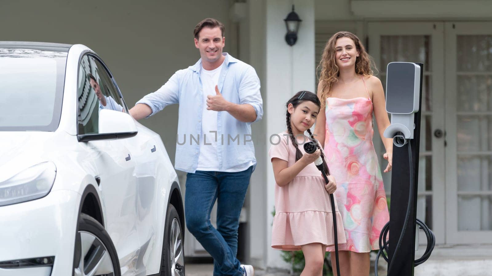 Modern family with environmental conscious recharging EV car with home EV charging station, Happy family with little young girl holding and pointing EV charger at camera. Synchronos