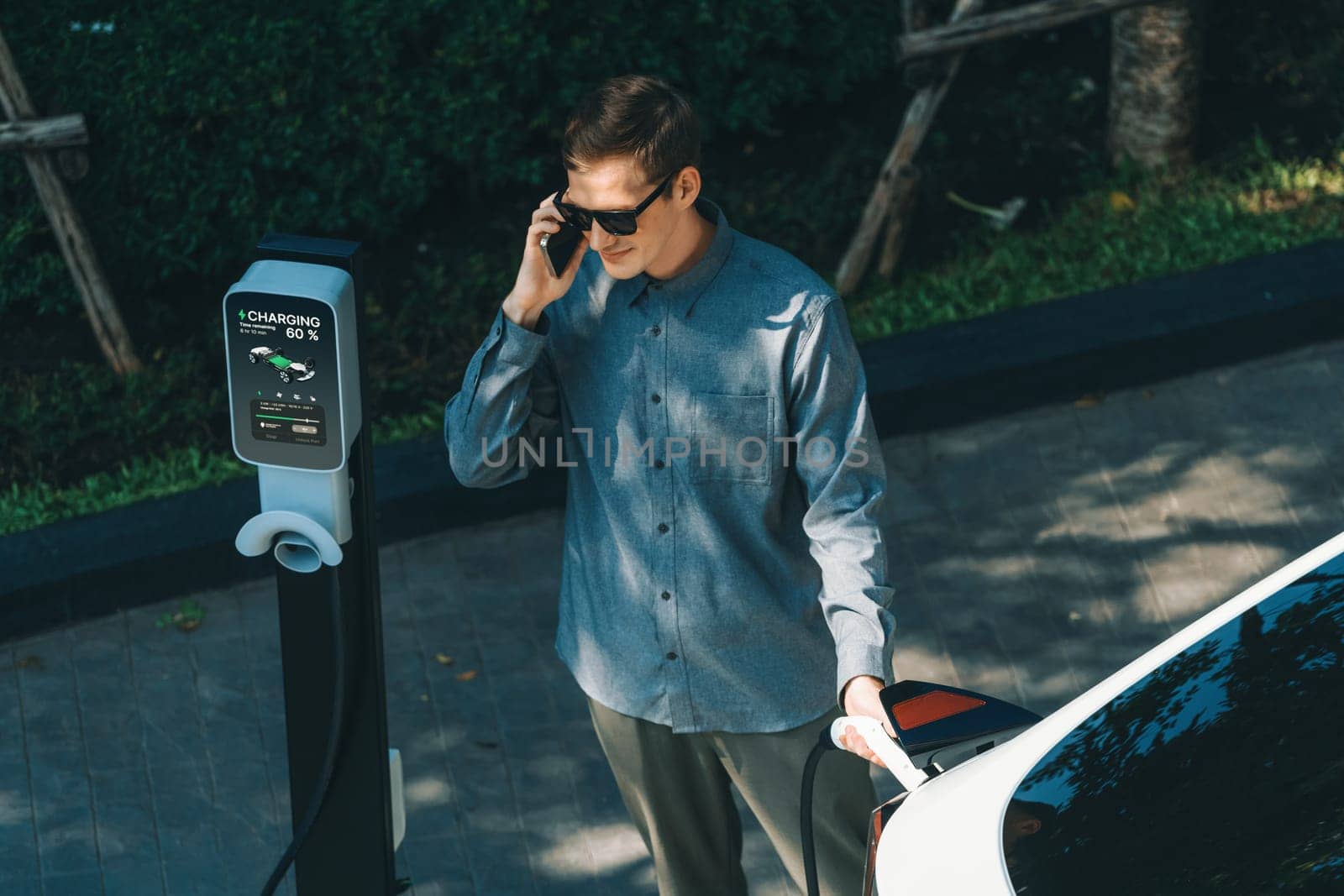 Young man travel with EV electric car charging in green sustainable city outdoor garden in summer shows urban sustainability lifestyle by green clean rechargeable energy of electric vehicle innards