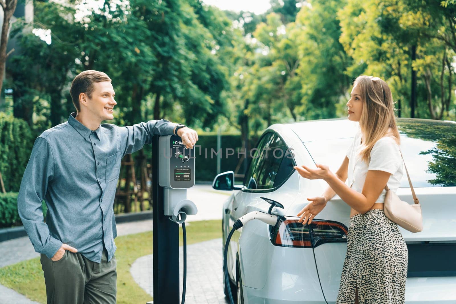 Young couple travel with EV electric car charging in green sustainable city outdoor garden in summer shows urban sustainability lifestyle by green clean rechargeable energy of electric vehicle innards