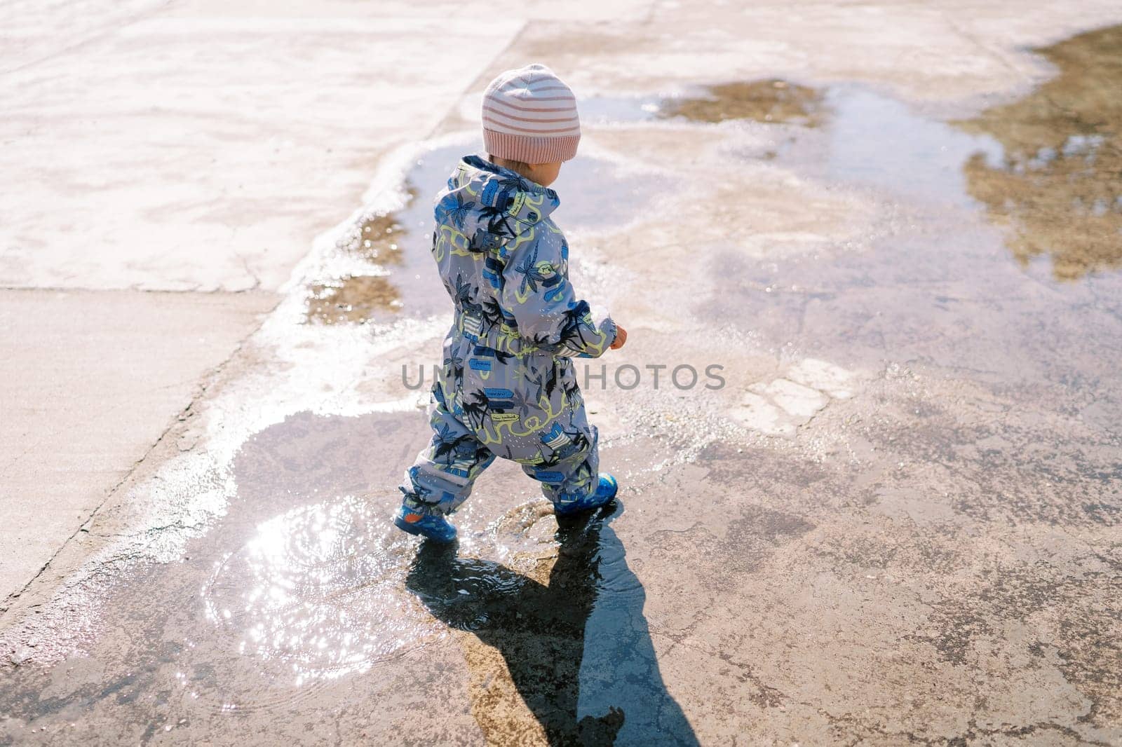 Little girl in rubber boots walks through a puddle on the pavement. Back view. High quality photo