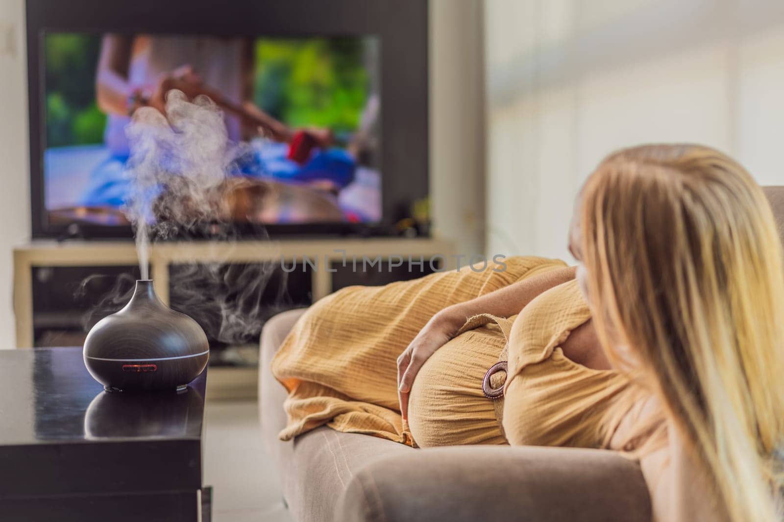 A blissful pregnant woman immerses in relaxation, savoring the soothing aroma from a diffuser while indulging in a calming TV video, embracing tranquility during her pregnant journey by galitskaya