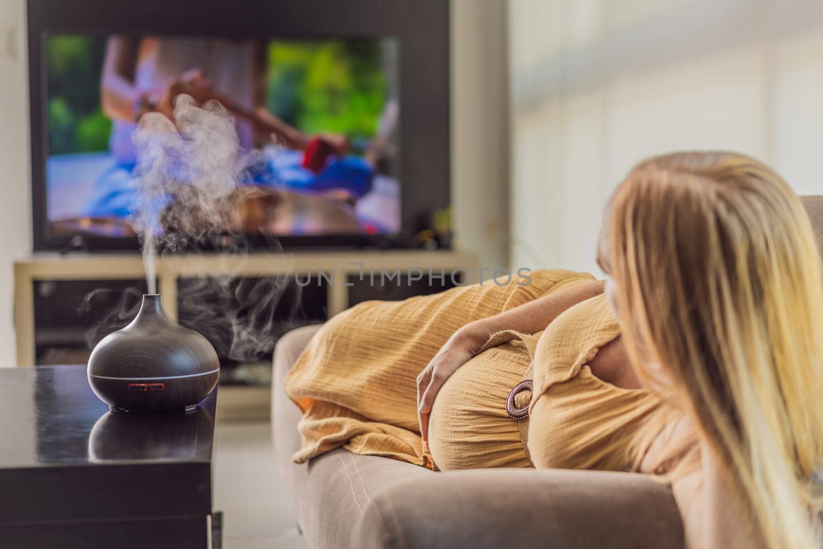 A blissful pregnant woman immerses in relaxation, savoring the soothing aroma from a diffuser while indulging in a calming TV video, embracing tranquility during her pregnant journey.