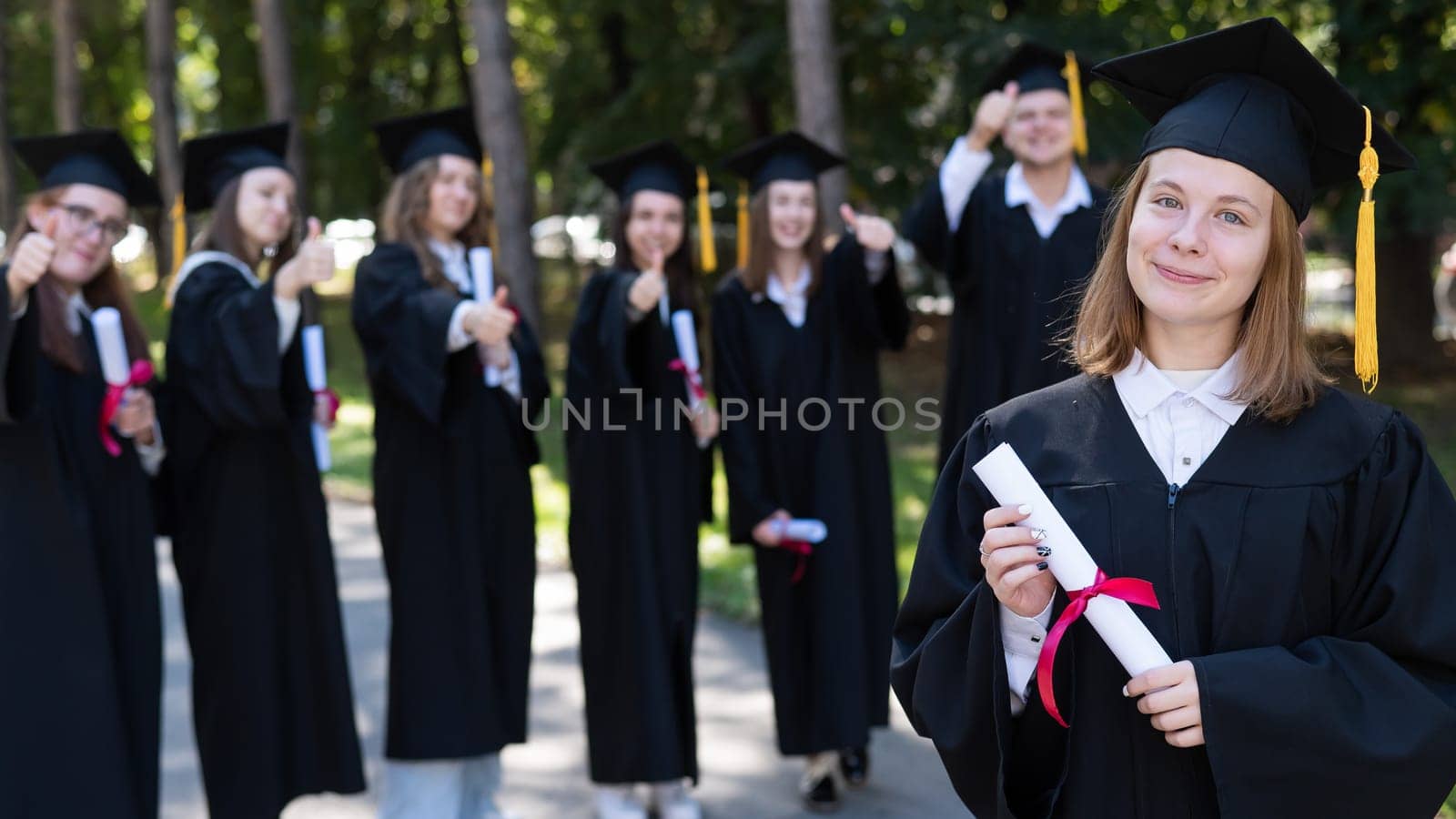 Group of happy students in graduation gowns outdoors. A young girl with a diploma in her hands in the foreground