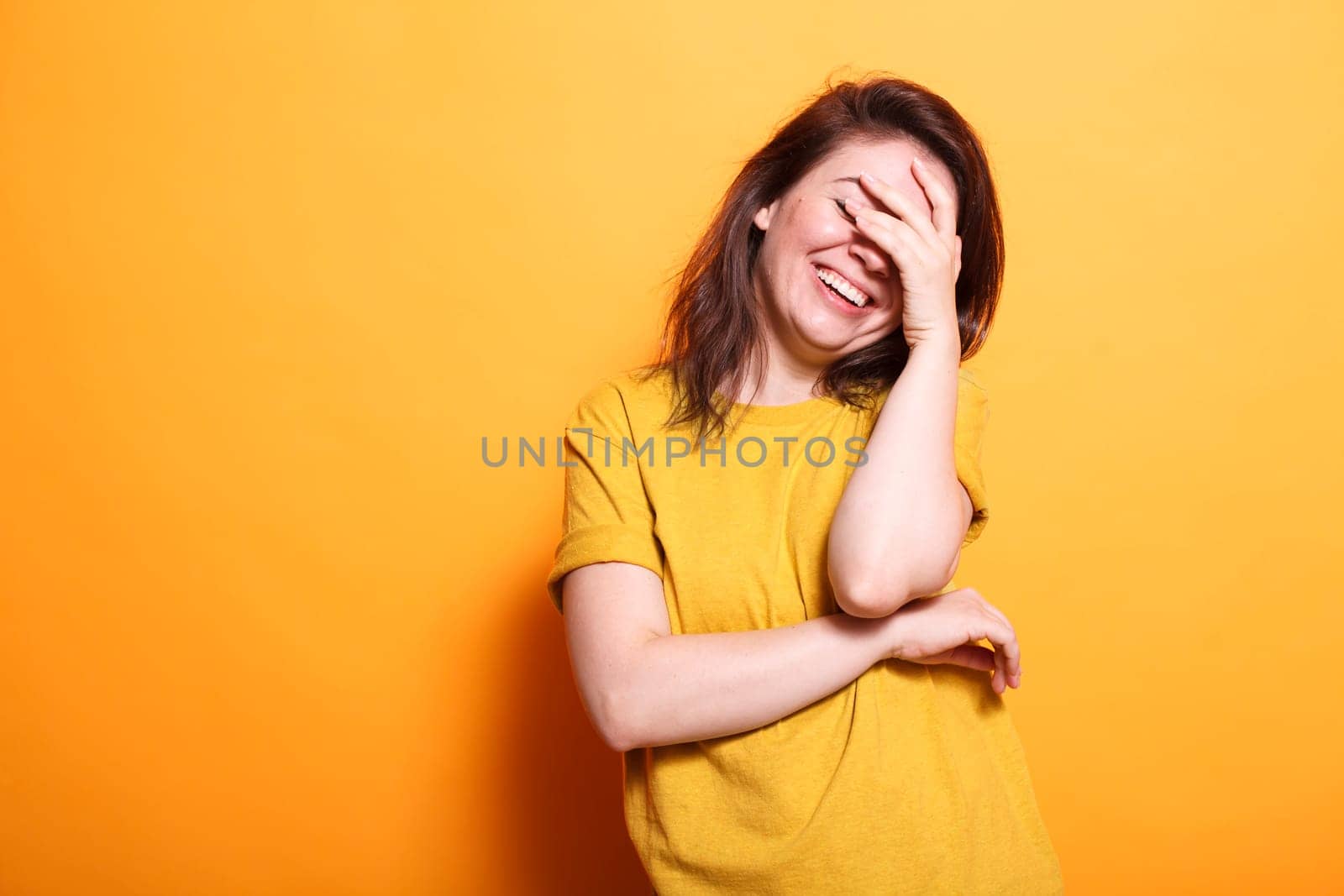 Portrait of beautiful brunette woman laughing while covering eyes and face. Enthusiastic lady using hand to hide from camera, feeling shy and happy standing over isolated background.