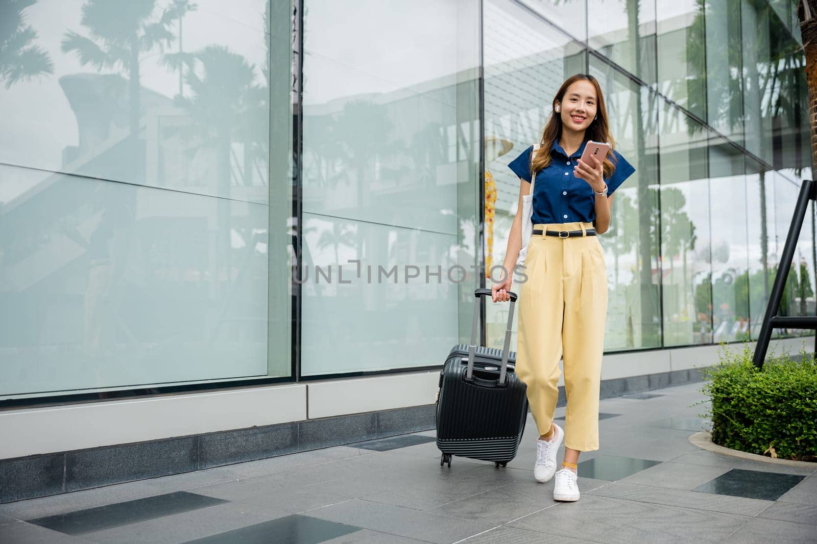 A stylish young black businesswoman on a business trip smiles while using her smartphone while walking down the city street with her luggage.