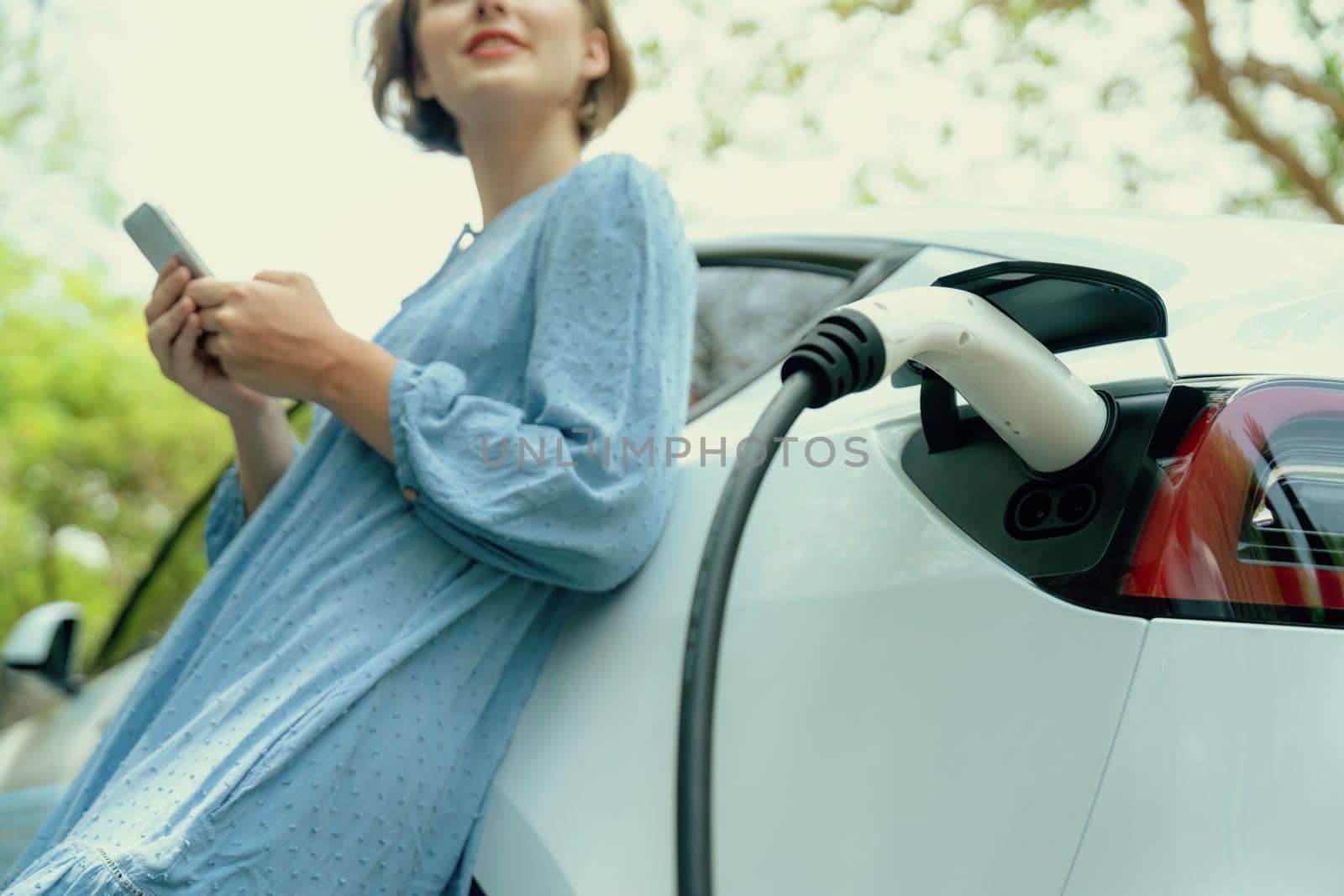 Focus EV charger from electric charging station recharging car on blurred background of beautiful young woman checking vehicle's battery from smartphone. Eco-friendly travel with EV car. Perpetual