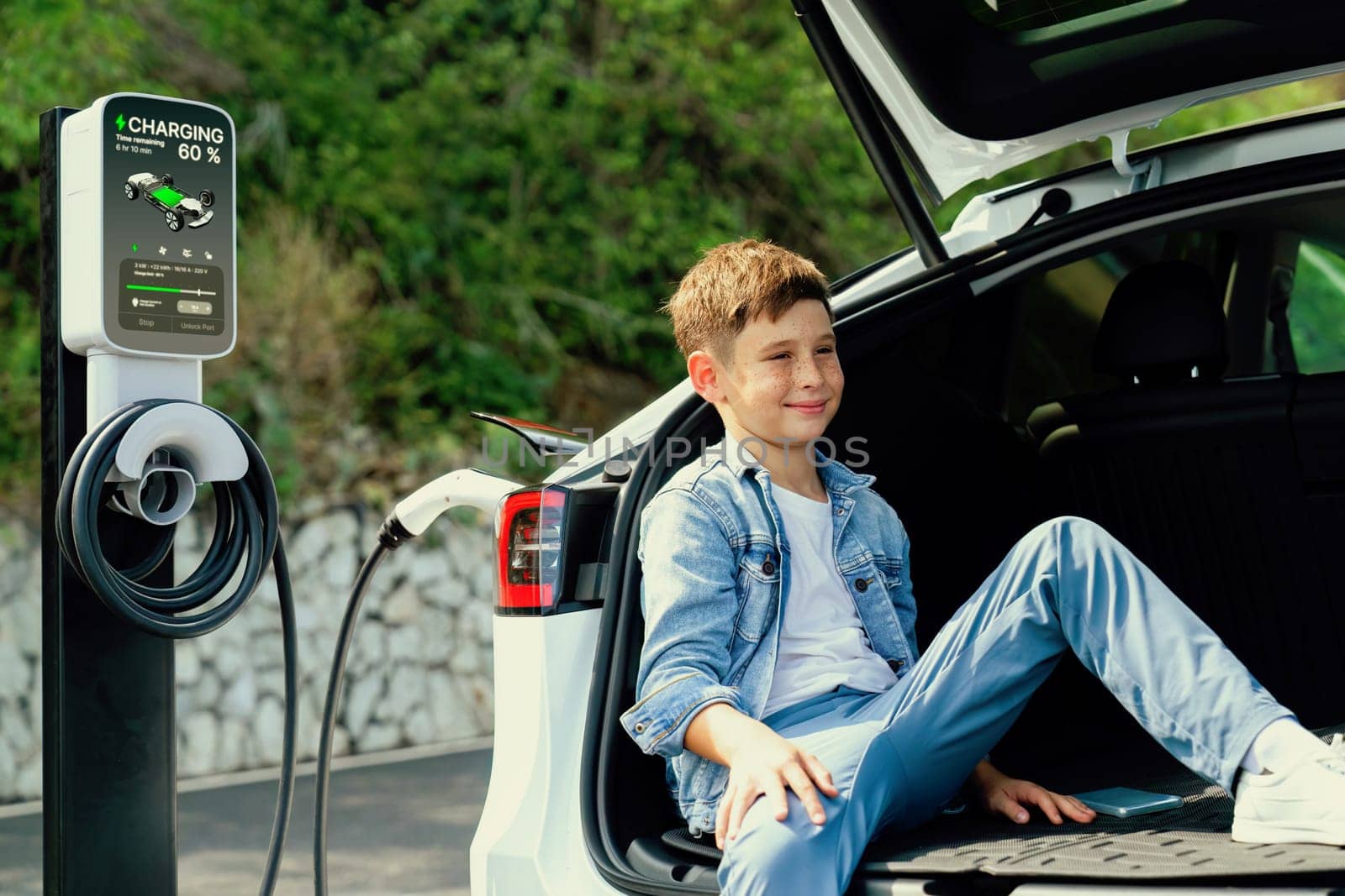 Little boy sitting on car trunk while recharging eco-friendly electric car from EV charging station. EV car road trip travel concept for alternative transportation powered sustainable energy.Perpetual