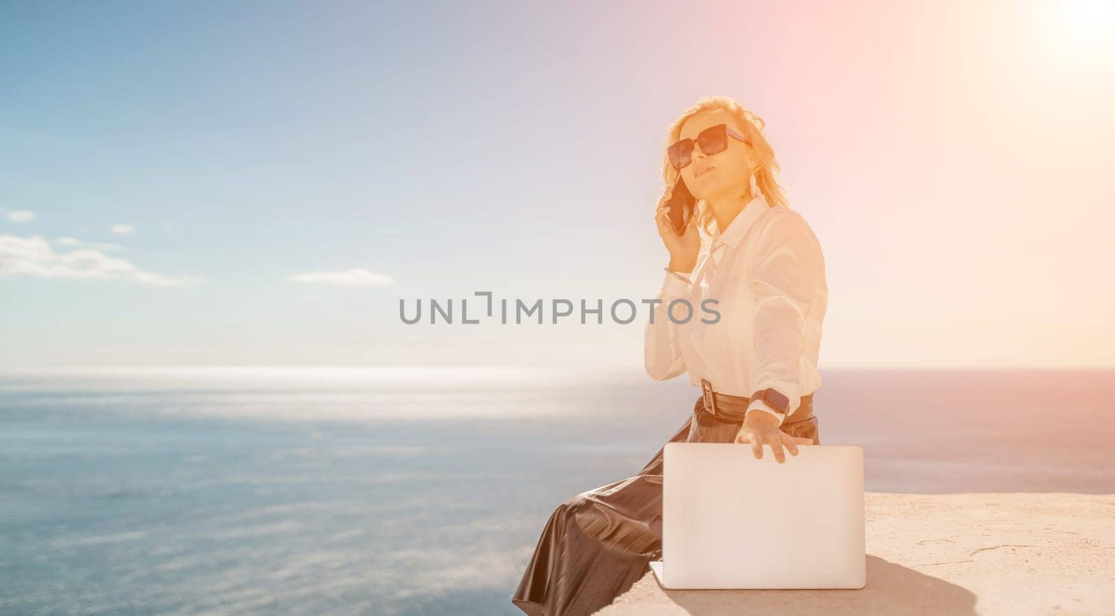 Freelance women sea working on a computer. Pretty middle aged woman with computer and phone outdoors with beautiful sea view. The concept of remote work. by Matiunina