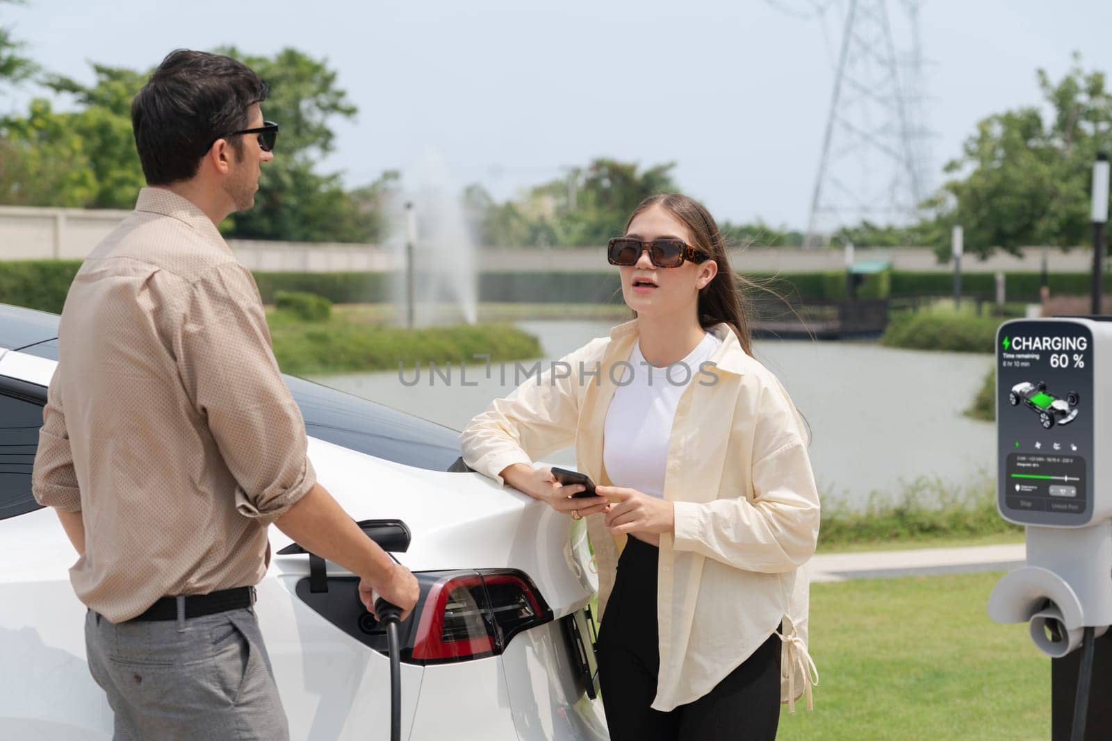 Young couple recharge electric car's battery from charging station in outdoor green city park in springtime. Rechargeable EV car for sustainable environmental friendly urban travel lifestyle.Expedient