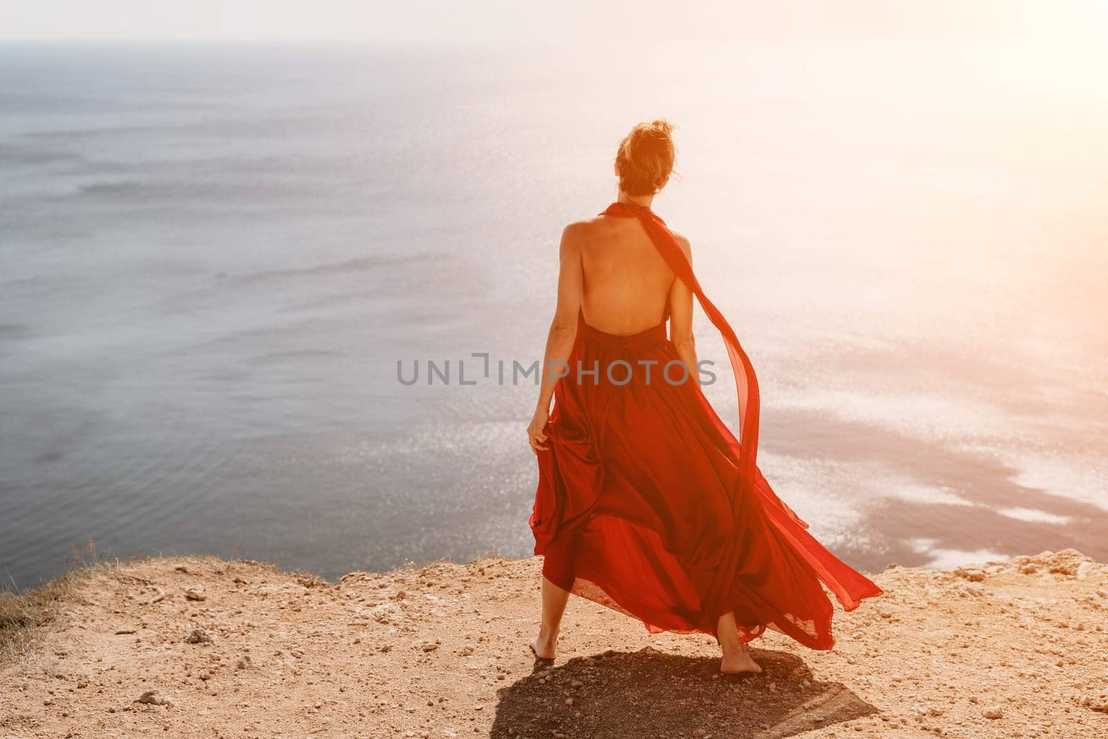 Side view a Young beautiful sensual woman in a red long dress posing on a rock high above the sea during sunrise. Girl on the nature on blue sky background. Fashion photo.