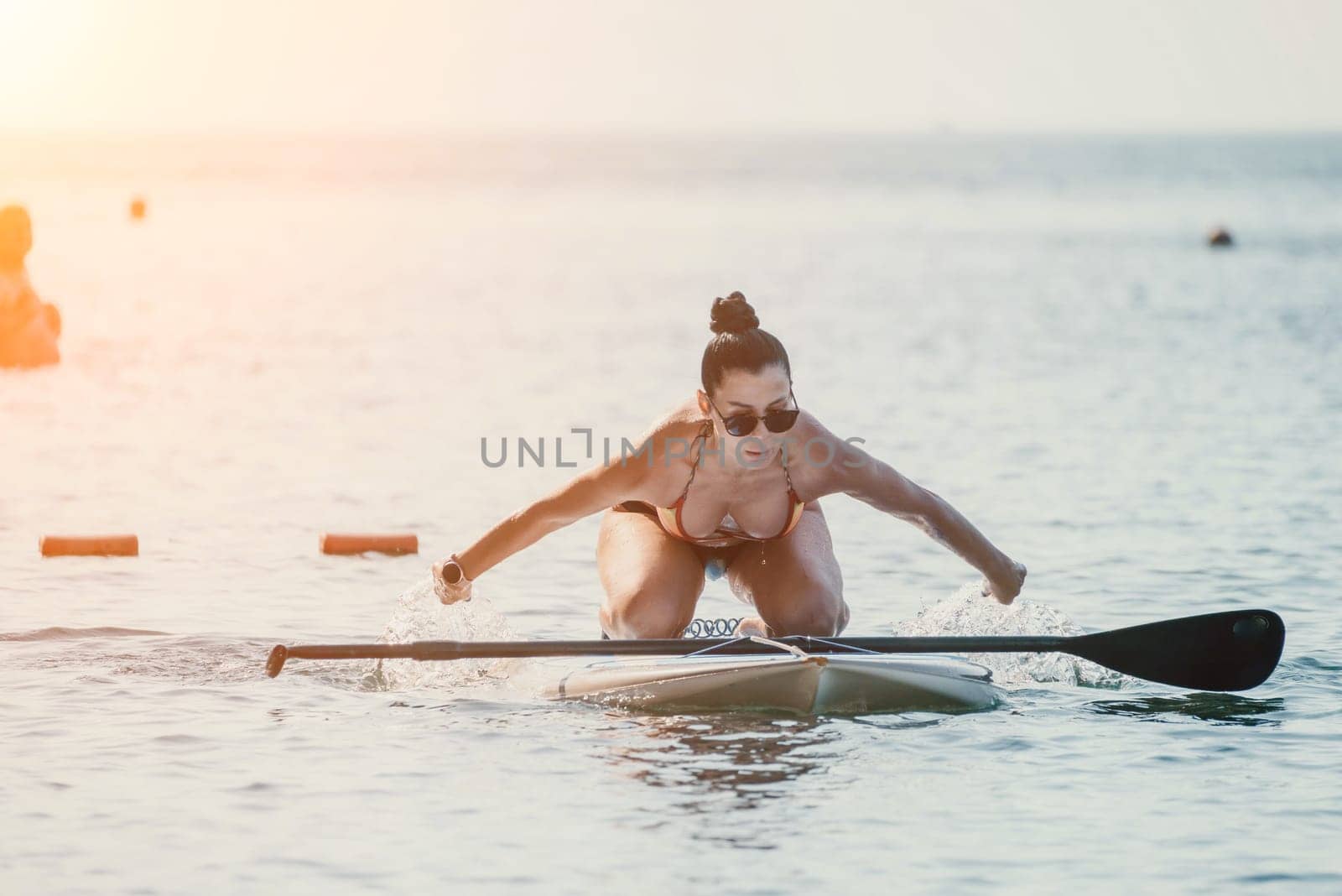 Sea woman sup. Silhouette of happy middle aged tanned woman in rainbow bikini, surfing on SUP board, confident paddling through water surface. Idyllic sunset. Active lifestyle at sea or river. by panophotograph