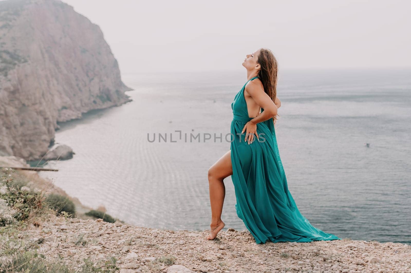 Woman travel portrait. Happy woman with long hair looking at camera and smiling. Close up portrait cute woman in a mint long dress posing on a volcanic rock high above the sea by panophotograph