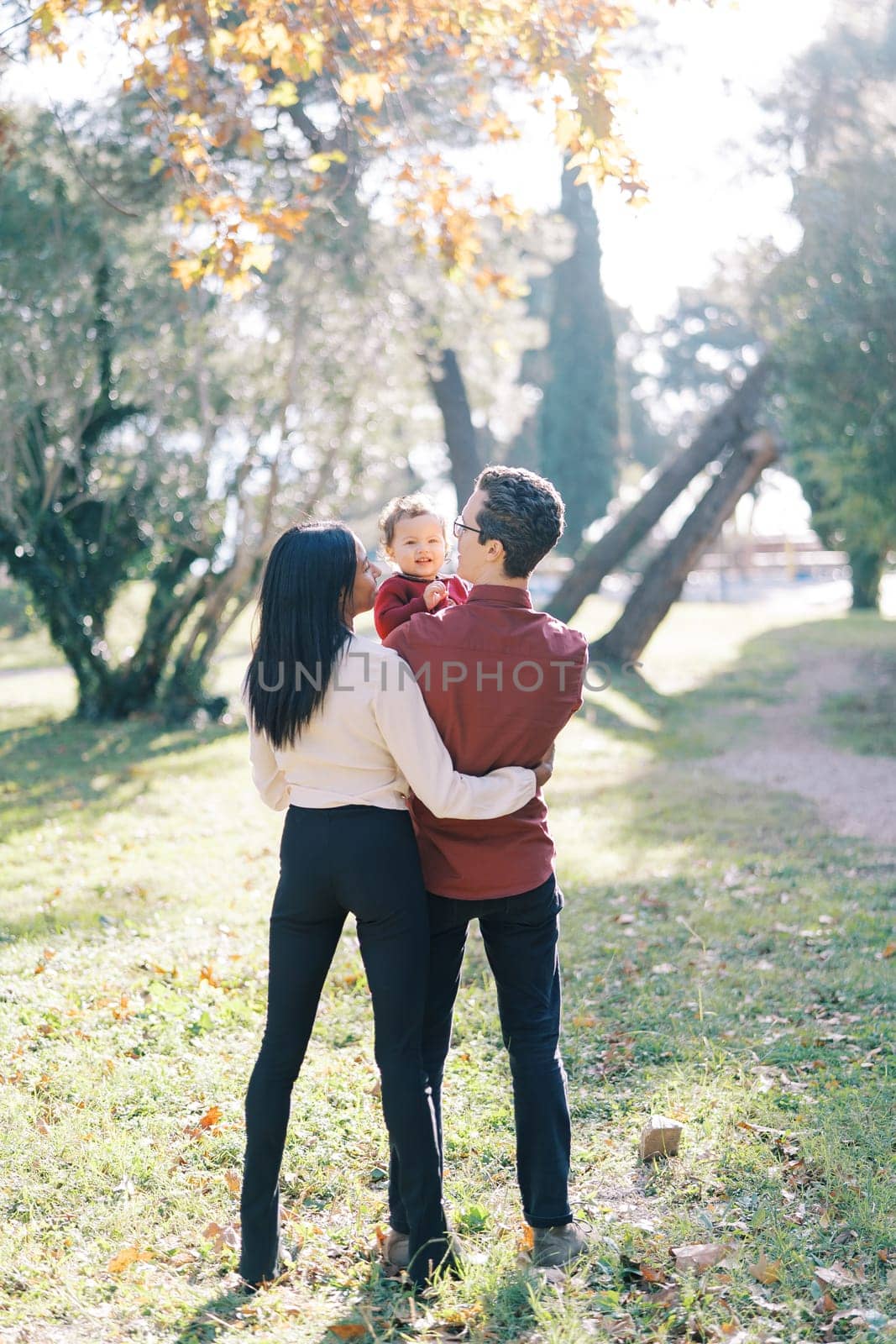 Mother hugs father with a little girl in his arms while standing in the park. Back view. High quality photo
