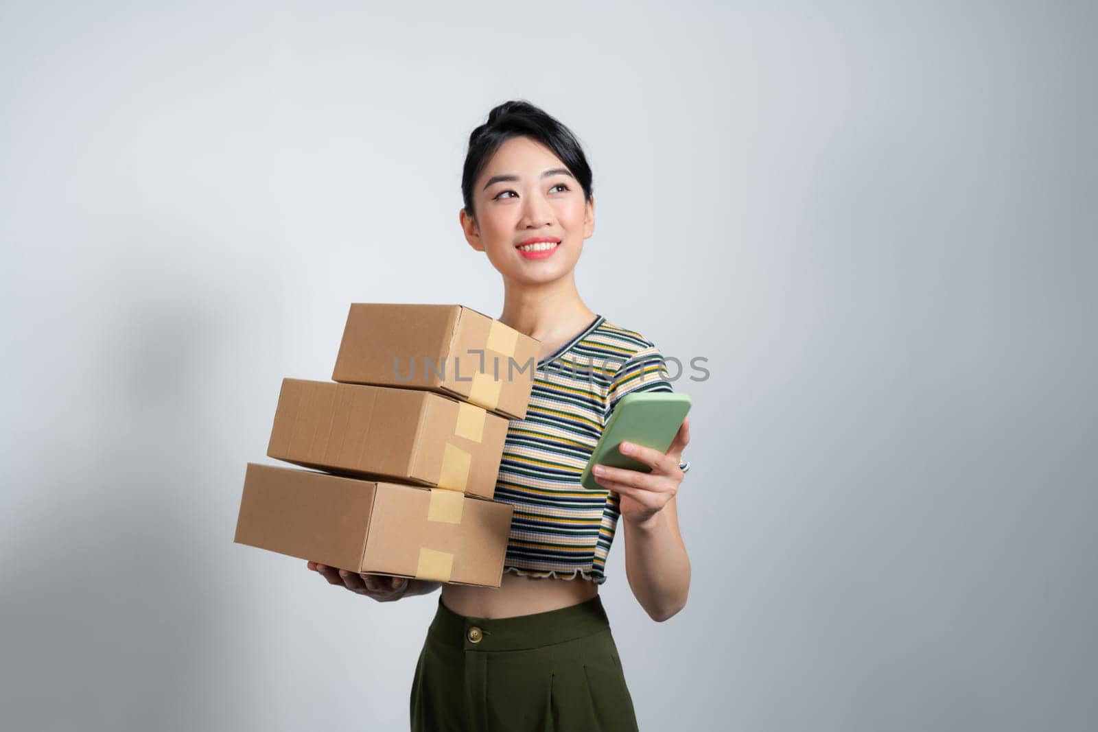 Young excited beautiful asian woman holding smartphone parcel cardboard standing on isolated white background