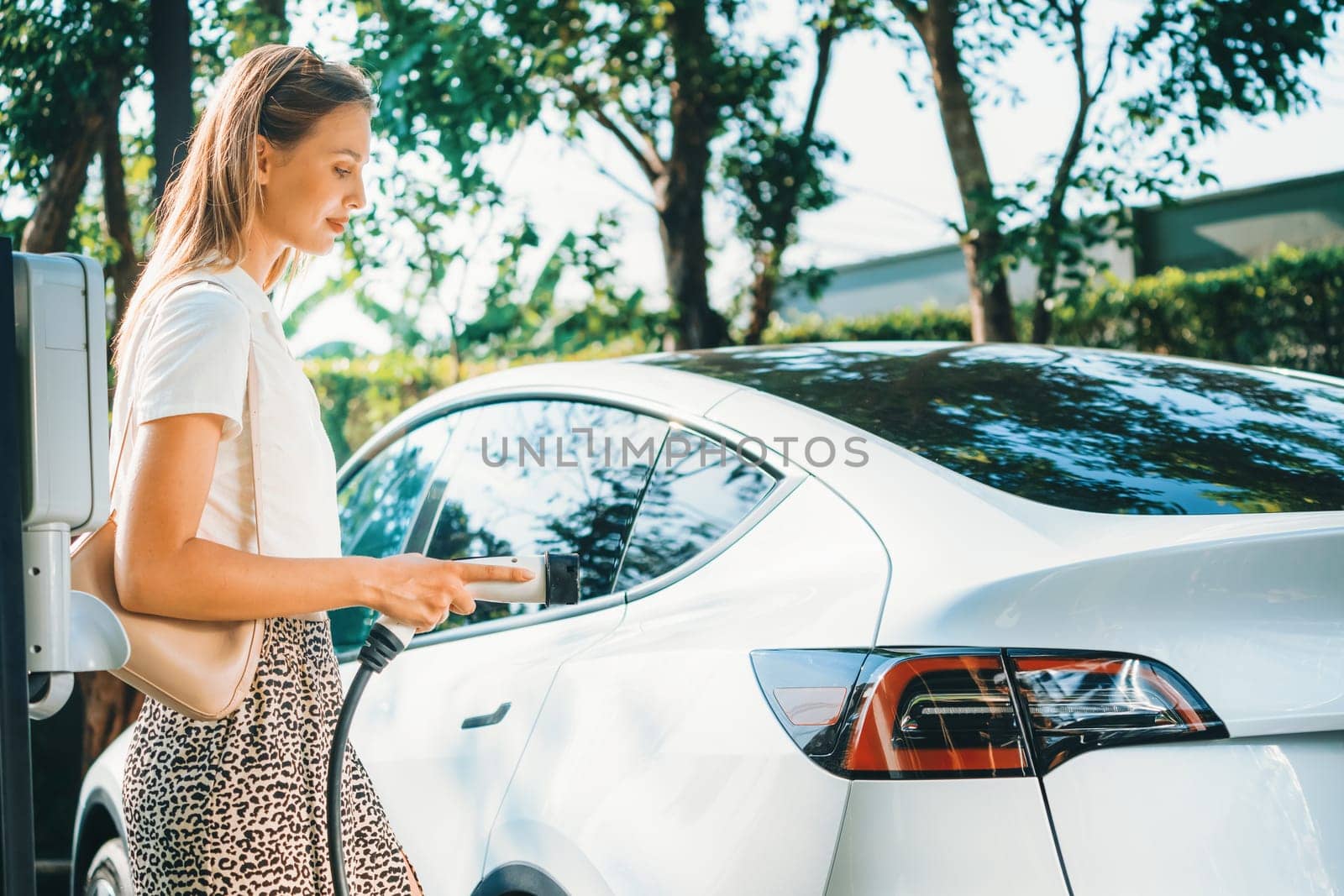 Young woman travel with EV electric car charging in green sustainable city outdoor garden in summer. Urban sustainability lifestyle by green clean rechargeable energy of electric BEV vehicle innards