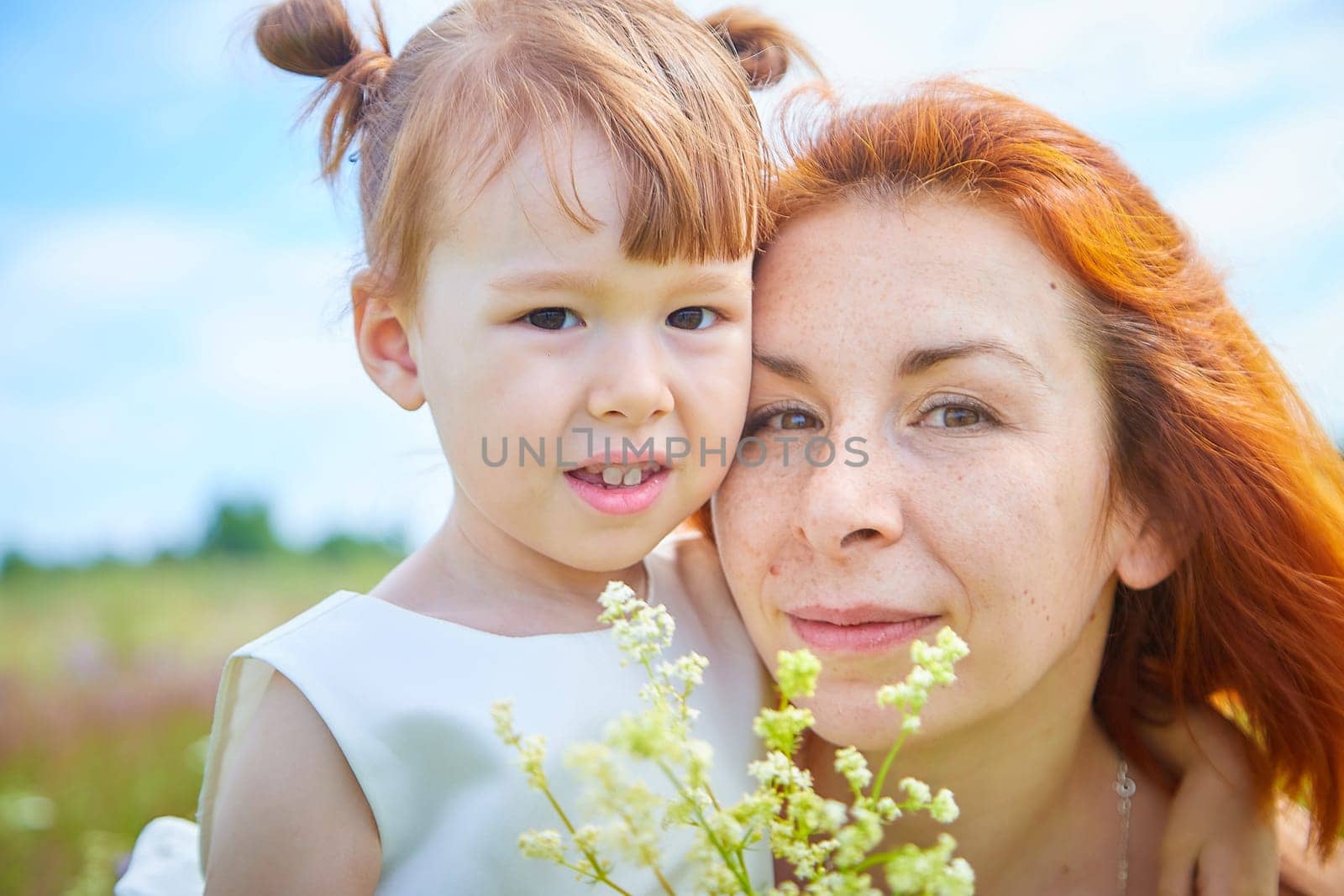 Happy female family with mother and daughter on green and yellow meadow full of grass and flower. Woman with red hair and blonde girl having fun, joy and hugs in sunny summer day. Concept family love by keleny