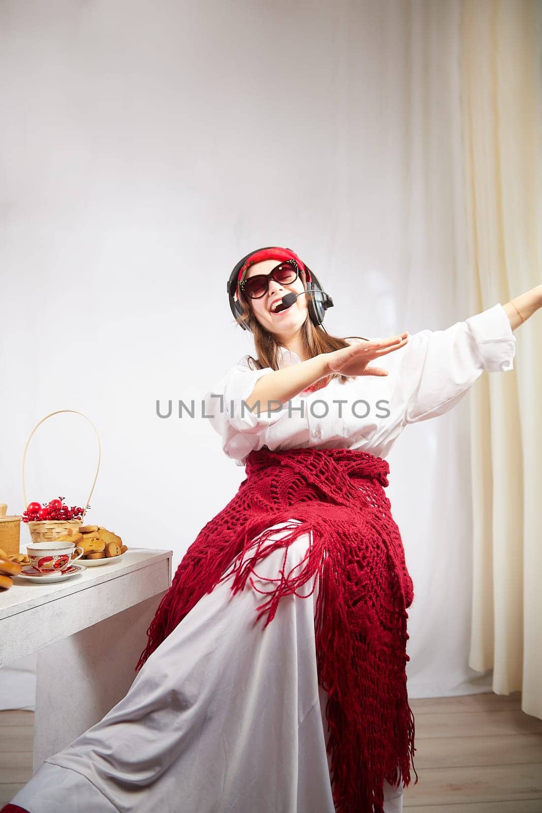 Girl in red scarf and large headphones with microphone having fun at table with delicious food for Orthodox holiday Maslenitsa or Easter. Funny Woman operator, Freelancer, blogger singing and dancing