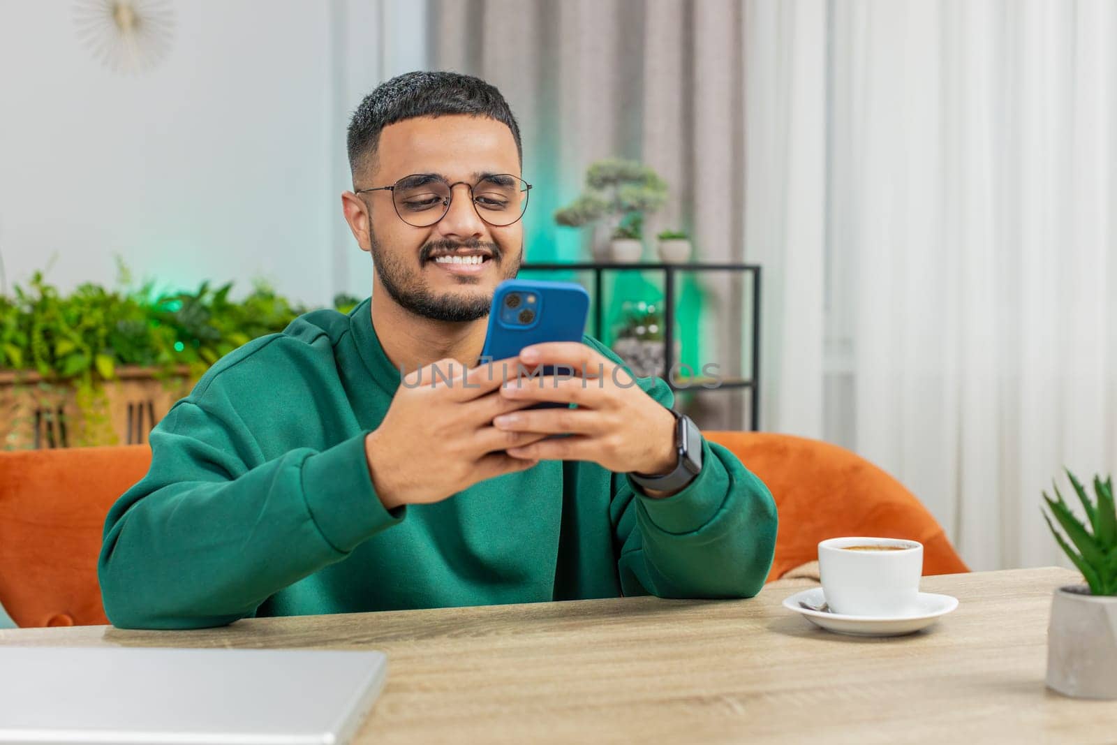 Indian Arabian young man sitting at table use smartphone share messages on social media application by efuror