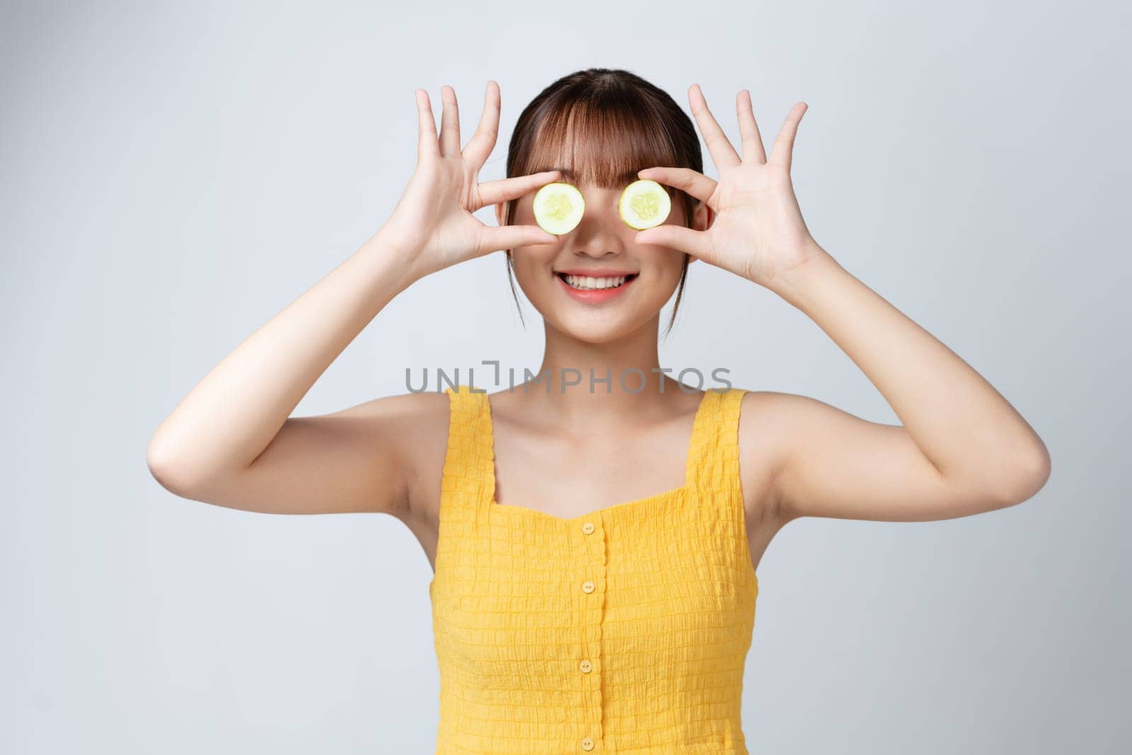 Beautiful and smiling young woman holding slice of cucumber in front of eyes on white background by makidotvn
