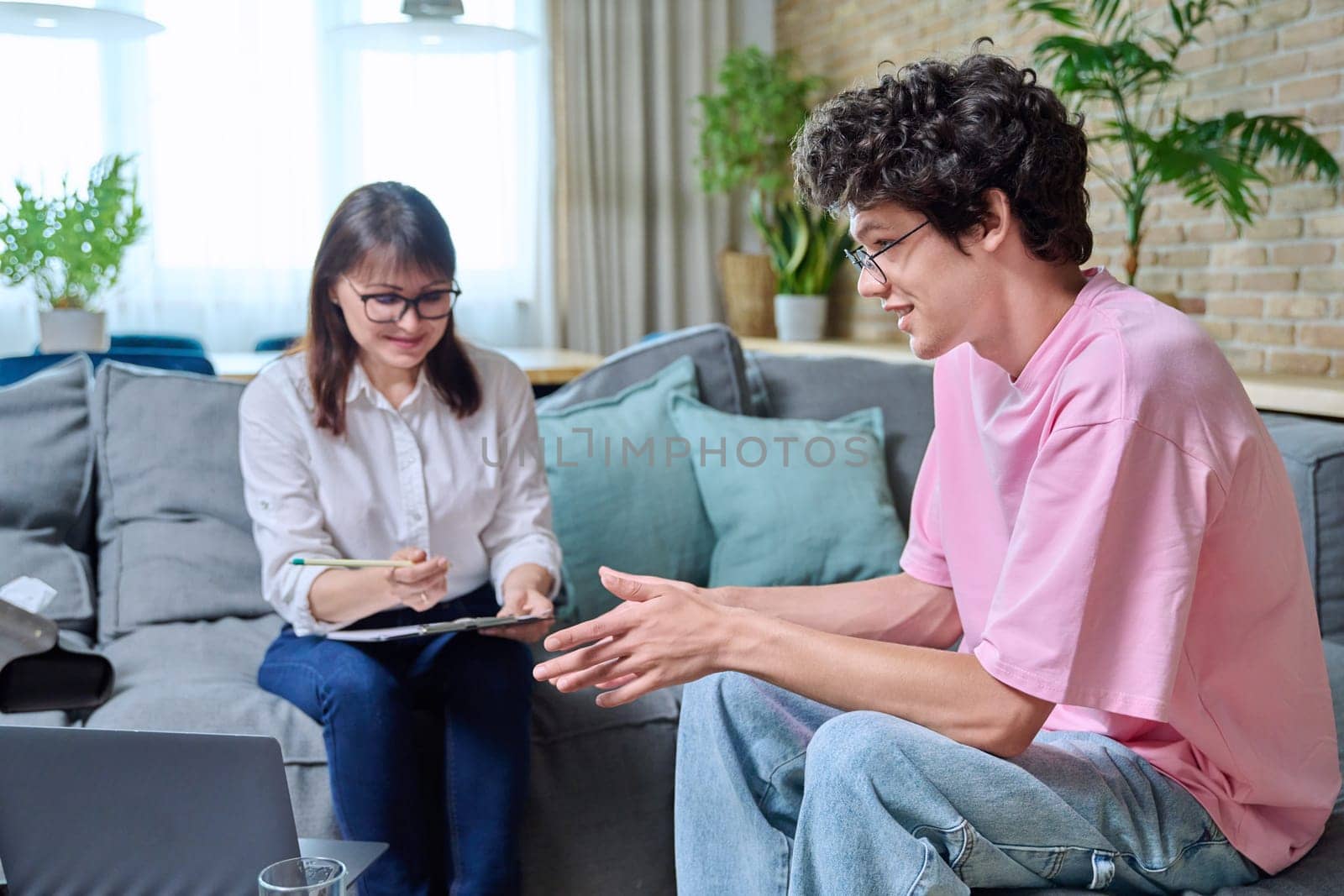 Young positive guy at therapy meeting with psychologist, patient and therapist sitting on sofa in office. Psychology, psychotherapy, treatment, mental health of youth concept